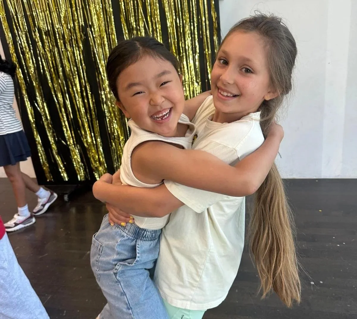 Two young girls hugging and smiling at the camera. One girl has dark hair and the other has long light brown hair. They are in a room with a gold tinsel backdrop.