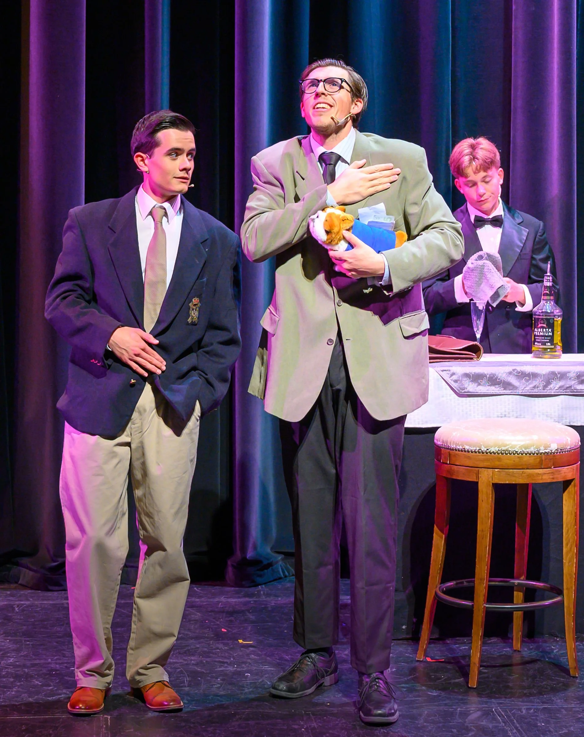 Three men on stage performing a comedy skit. The man in the center, wearing glasses and a beige blazer, is holding a toy dog and has his hand on his chest. The man on the left is dressed in a dark blazer and light pants, showing an amused expression.