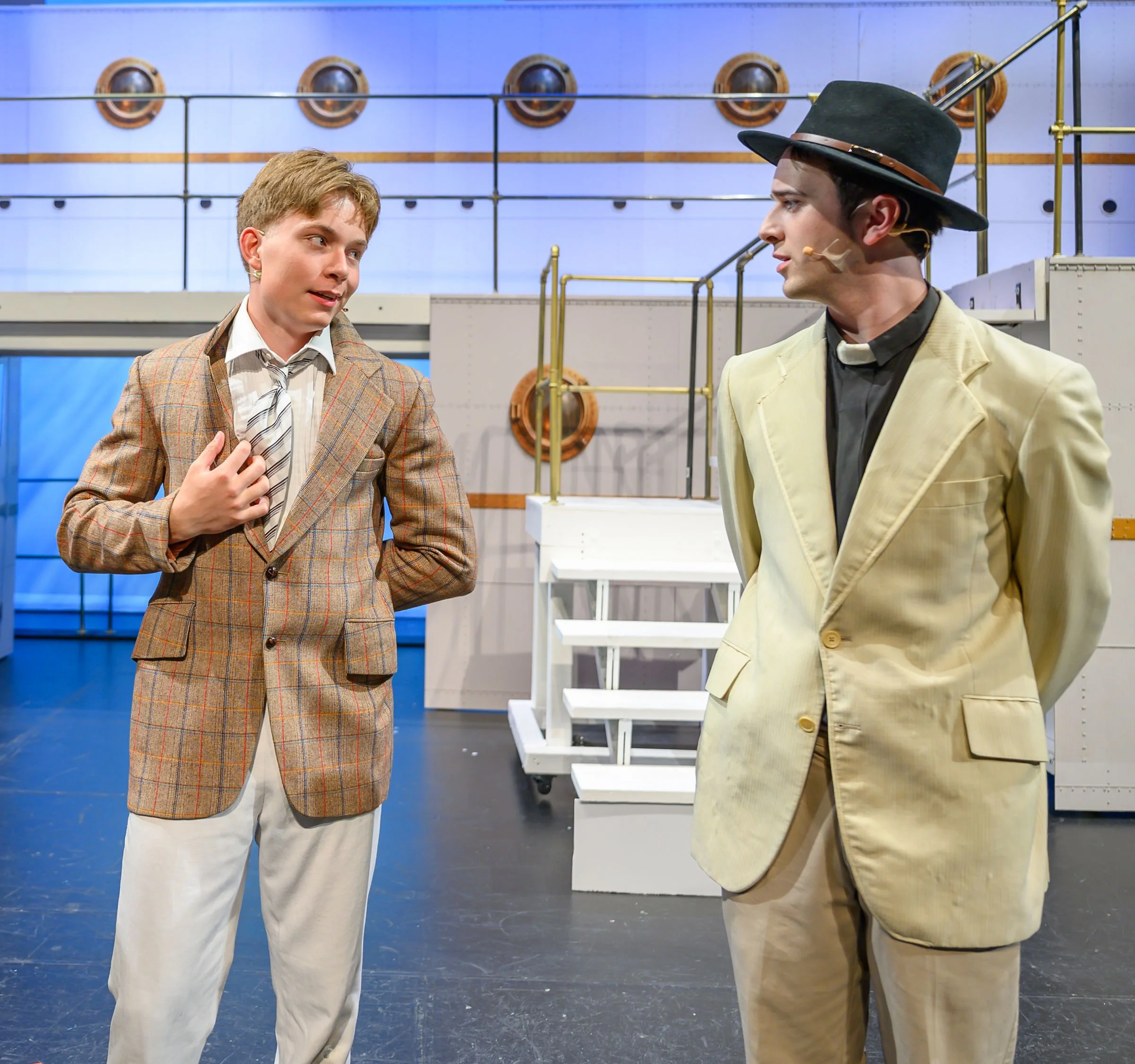 Two men dressed in vintage or theatrical costumes are conversing indoors. The man on the left wears a brown plaid blazer with a white shirt and a striped tie, and the man on the right wears a yellow blazer with a black shirt and a large black hat. Th