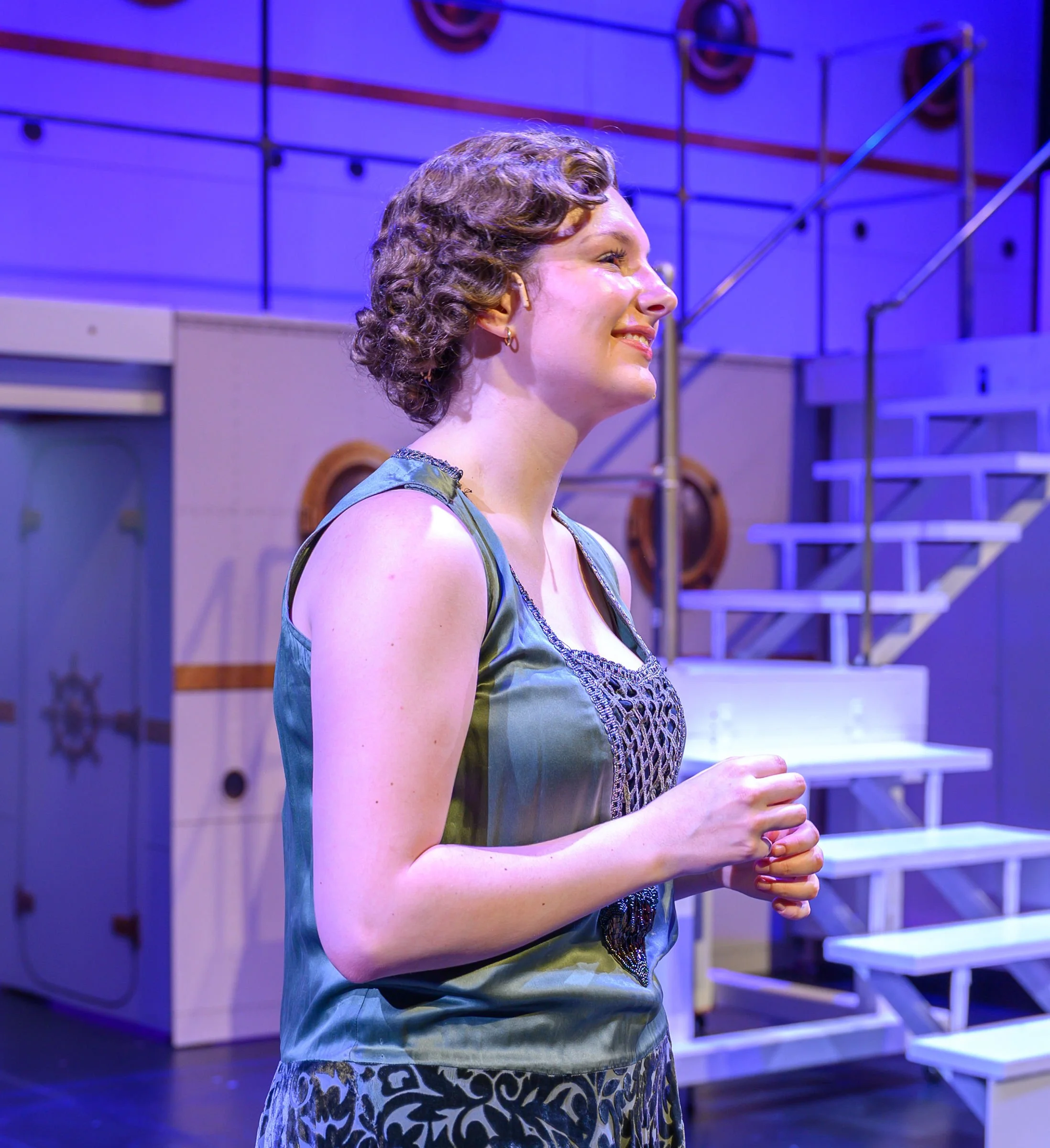 A woman with short curly hair and earrings, smiling and standing in front of lockers with a staircase in the background, illuminated by blue and purple lighting.