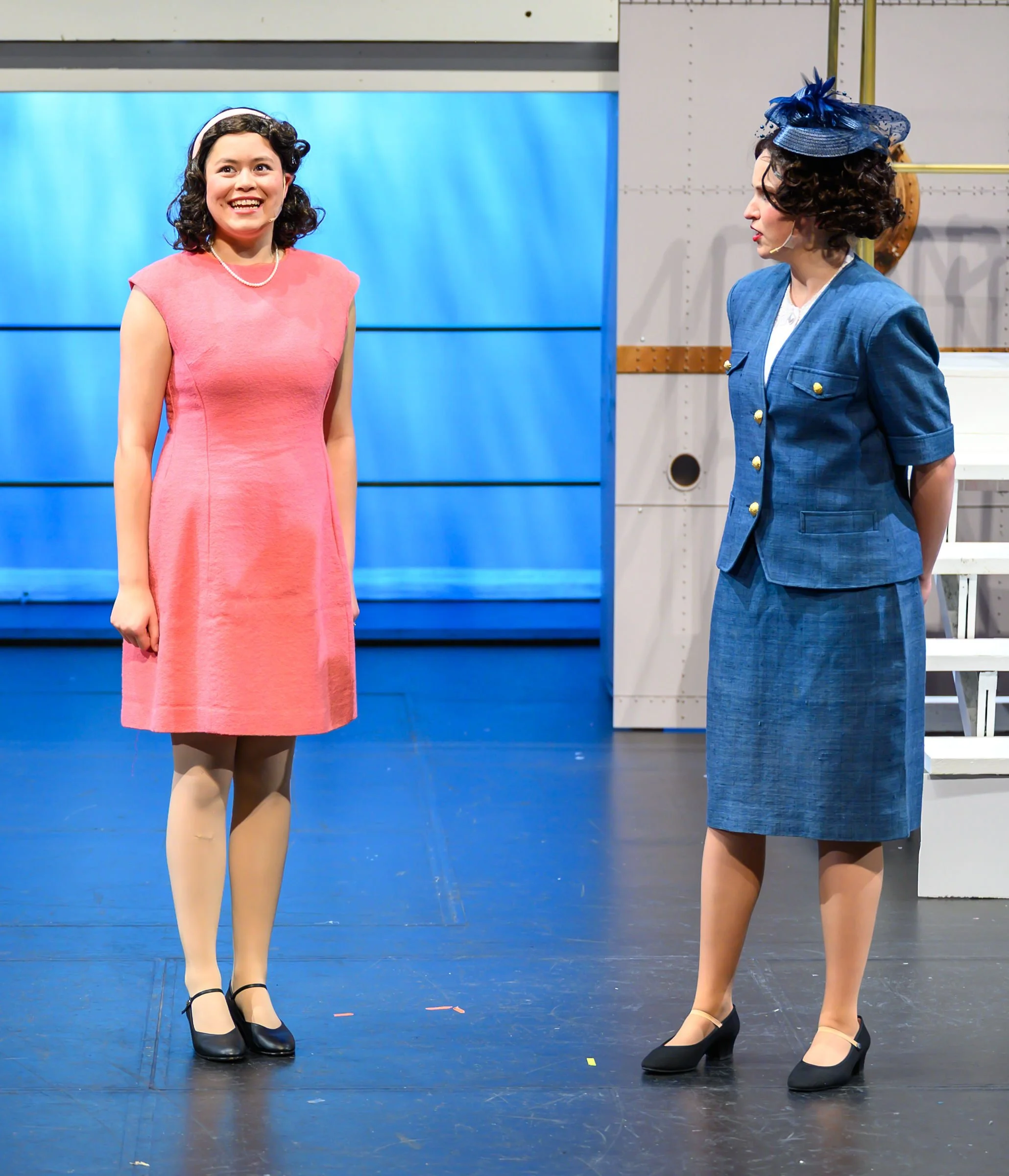 Two women dressed in vintage-style clothing and heels, standing on a stage with a blue backdrop. One woman is smiling while the other is turned to the side, both appear to be engaged in conversation or performance.