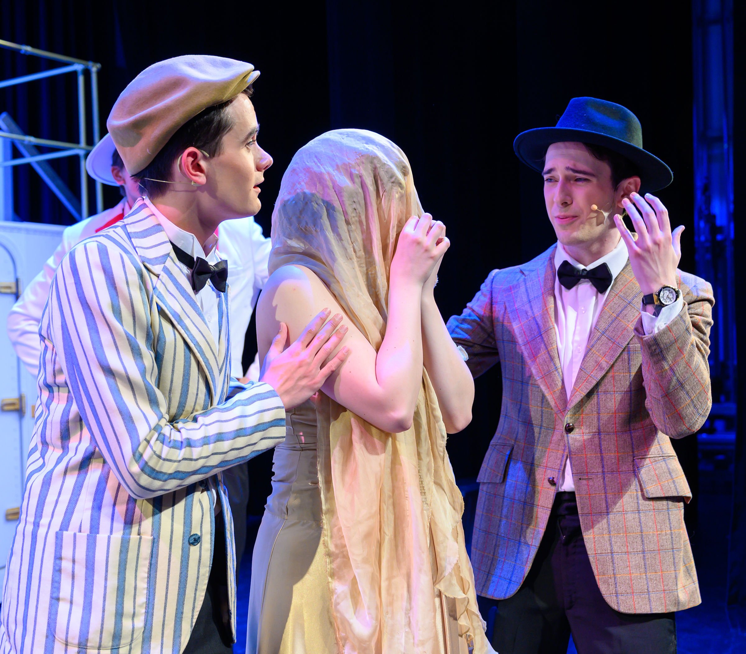 Three performers on stage, two men in colorful suits with bow ties and hats, one woman with a paper bag over her head, all engaging in a theatrical performance.