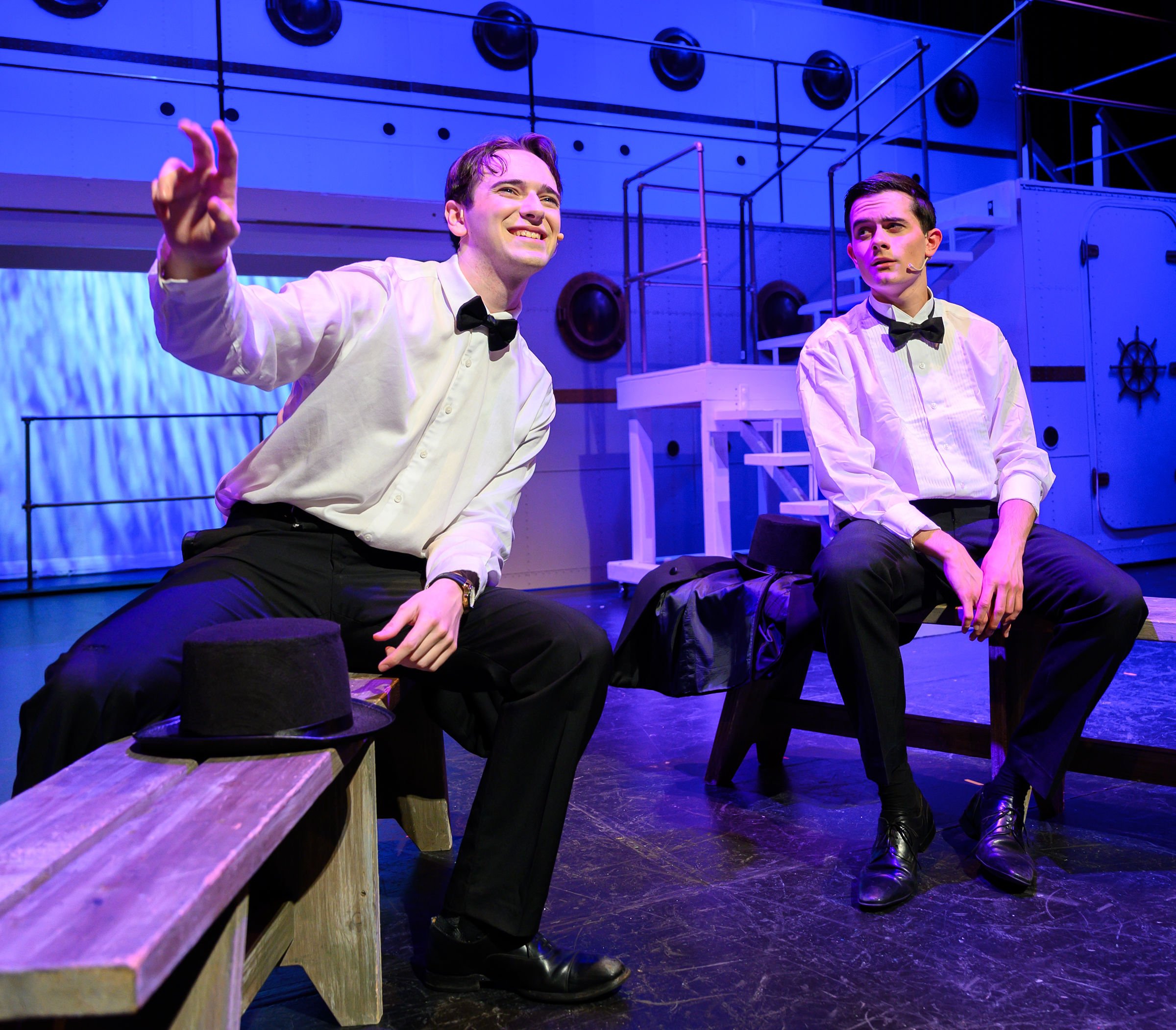 Two men dressed as waiters with bow ties sitting on a stage in a theater, with a backdrop of blue and purple lighting, wooden benches, and stage equipment.