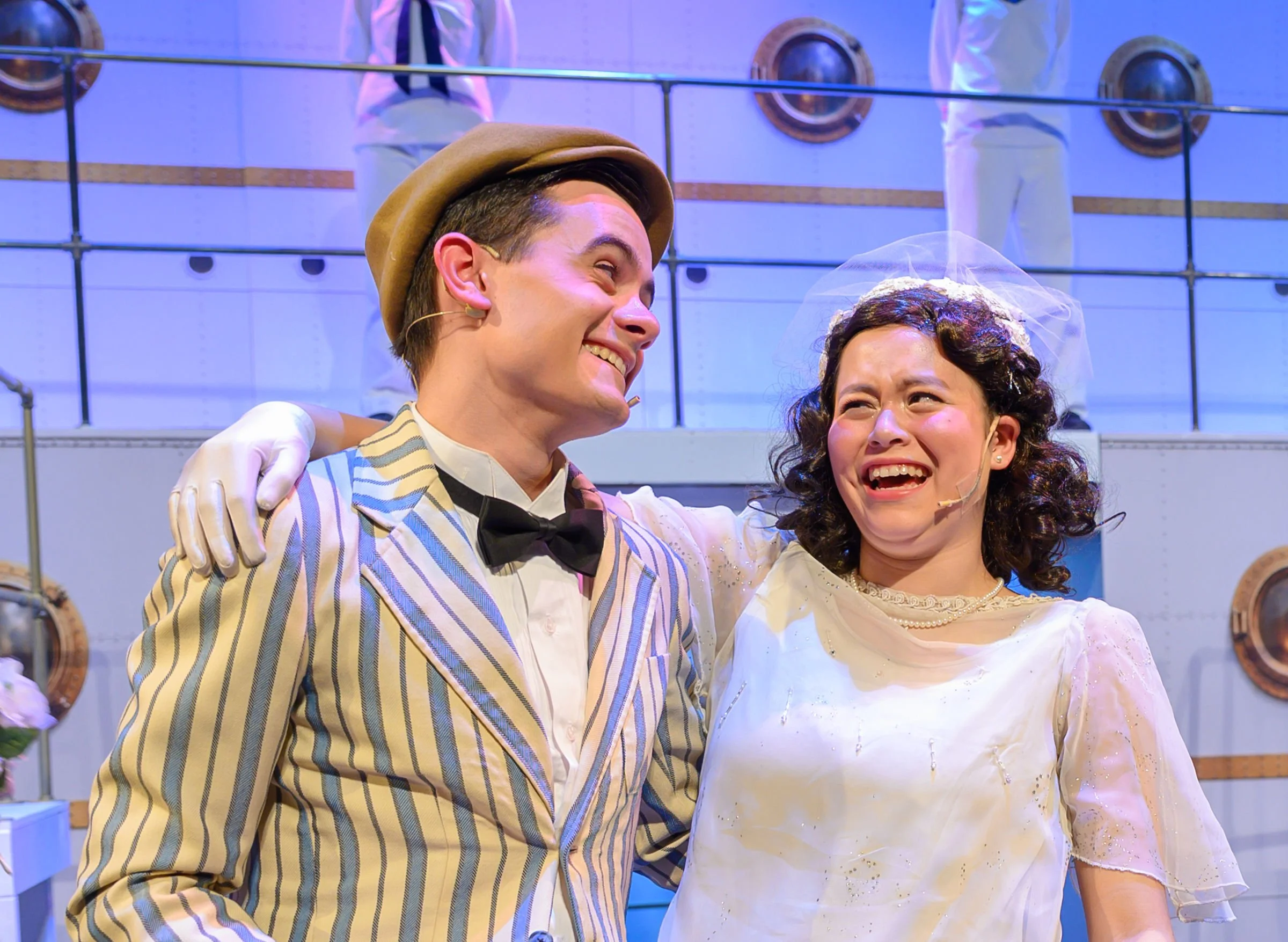 Two actors in costumes smile and laugh while embracing each other, with a background of stage scenery including wood paneling and circular portholes.