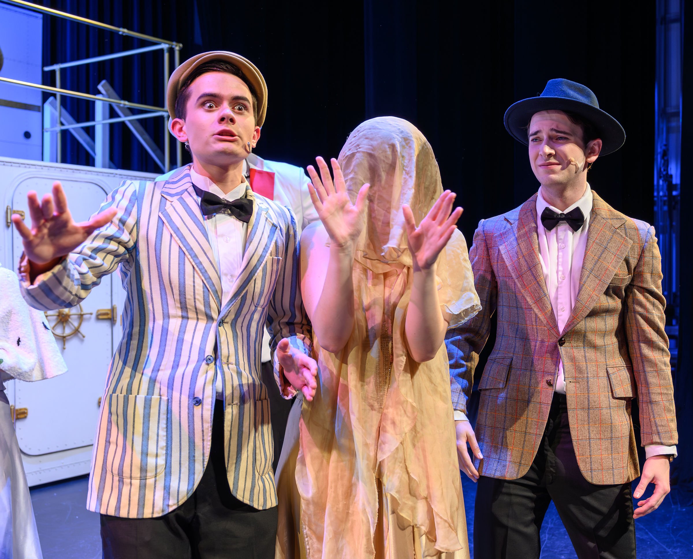 Three men and a woman are on stage during a theatrical performance. The man on the left, with dark hair, is wearing a striped suit and a boutonniere, and is expressive with his hands raised. The middle person, with their face covered in a beige cloth