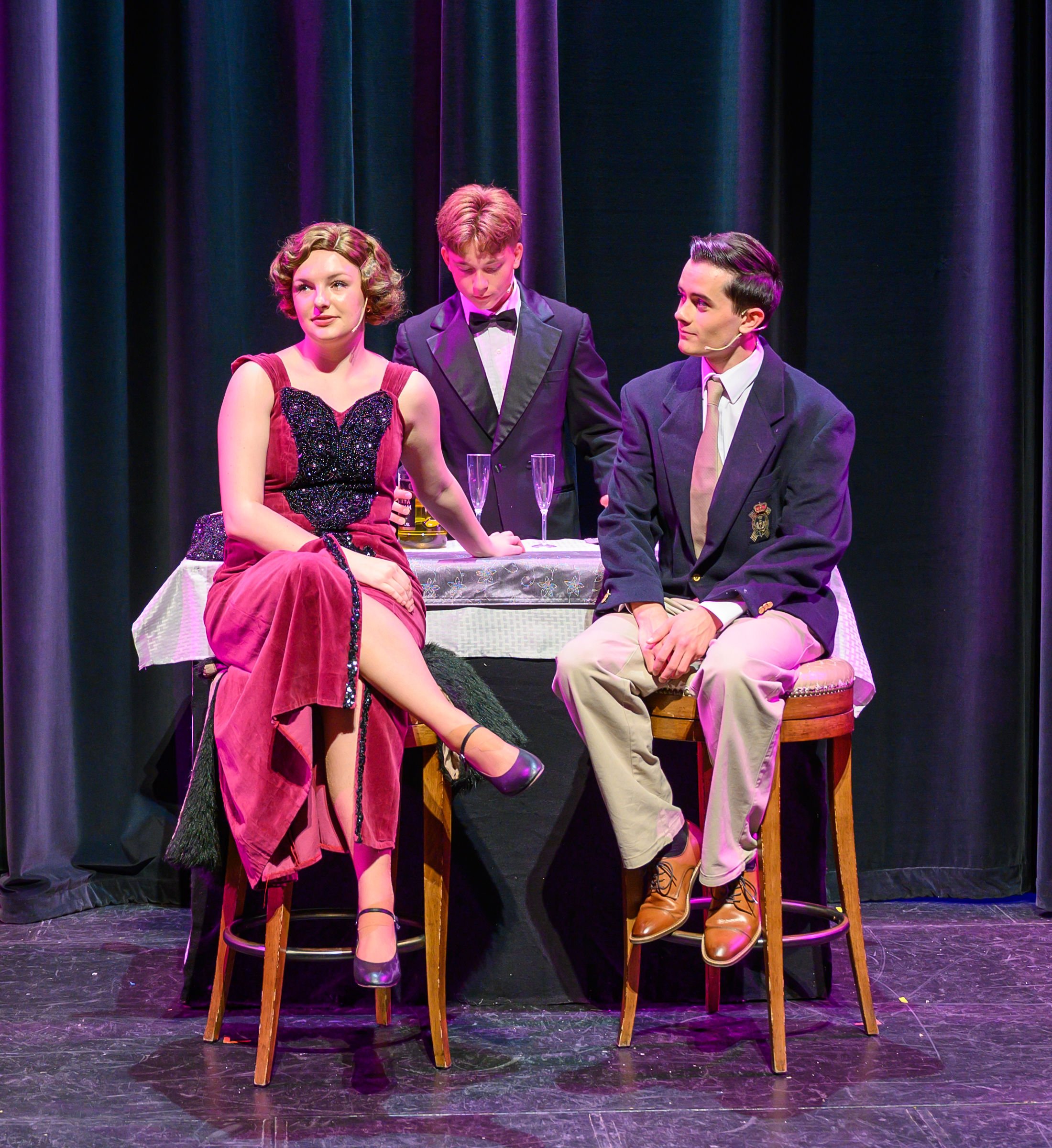 Three young actors on stage, two men and one woman, dressed in vintage formal attire, sitting on bar stools behind a table with champagne flutes, with theatrical curtains in the background.