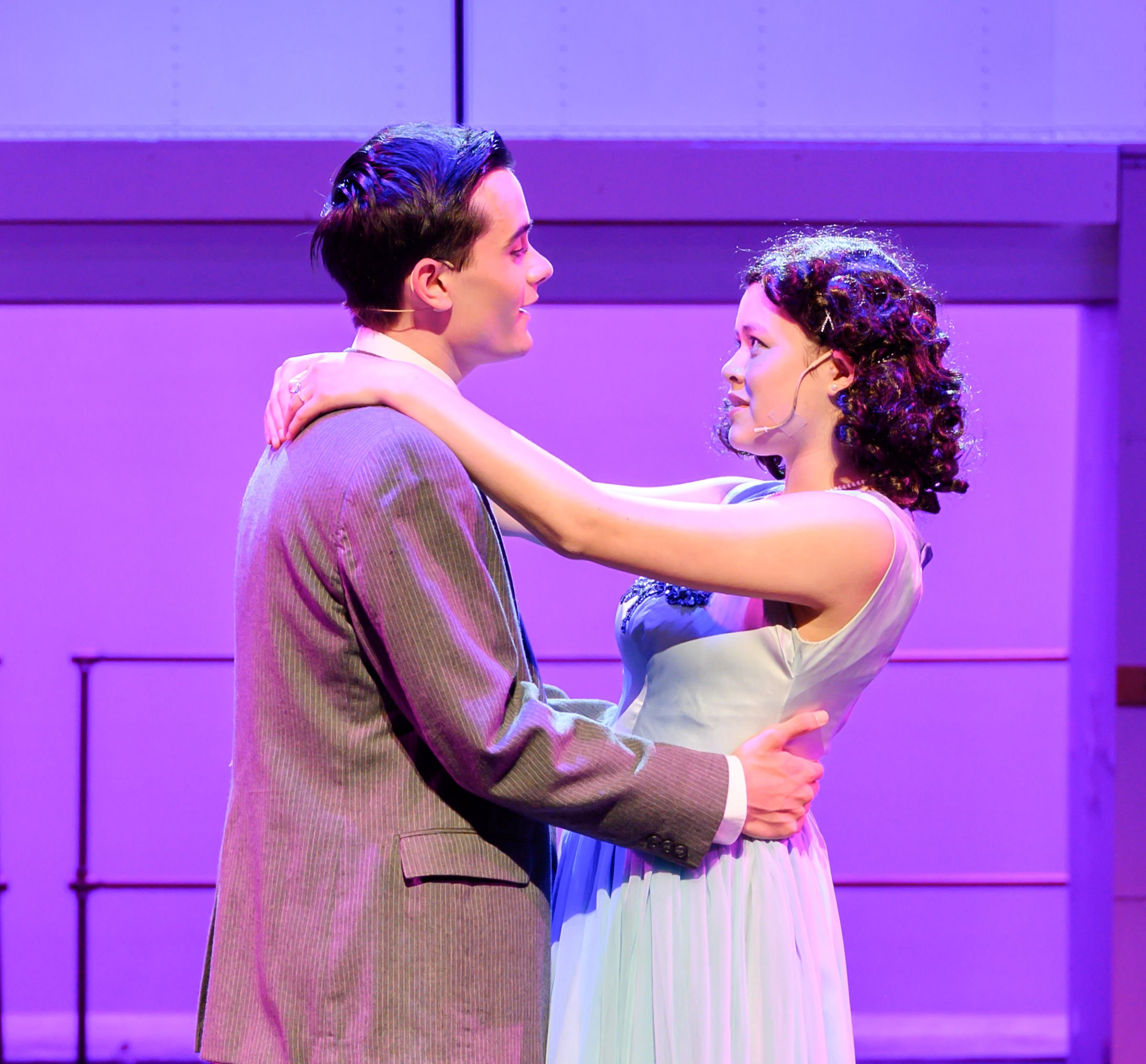A man and a woman on stage, facing each other closely, with the man smiling and holding her waist, and the woman looking up at him, with purple lighting and a simple background.