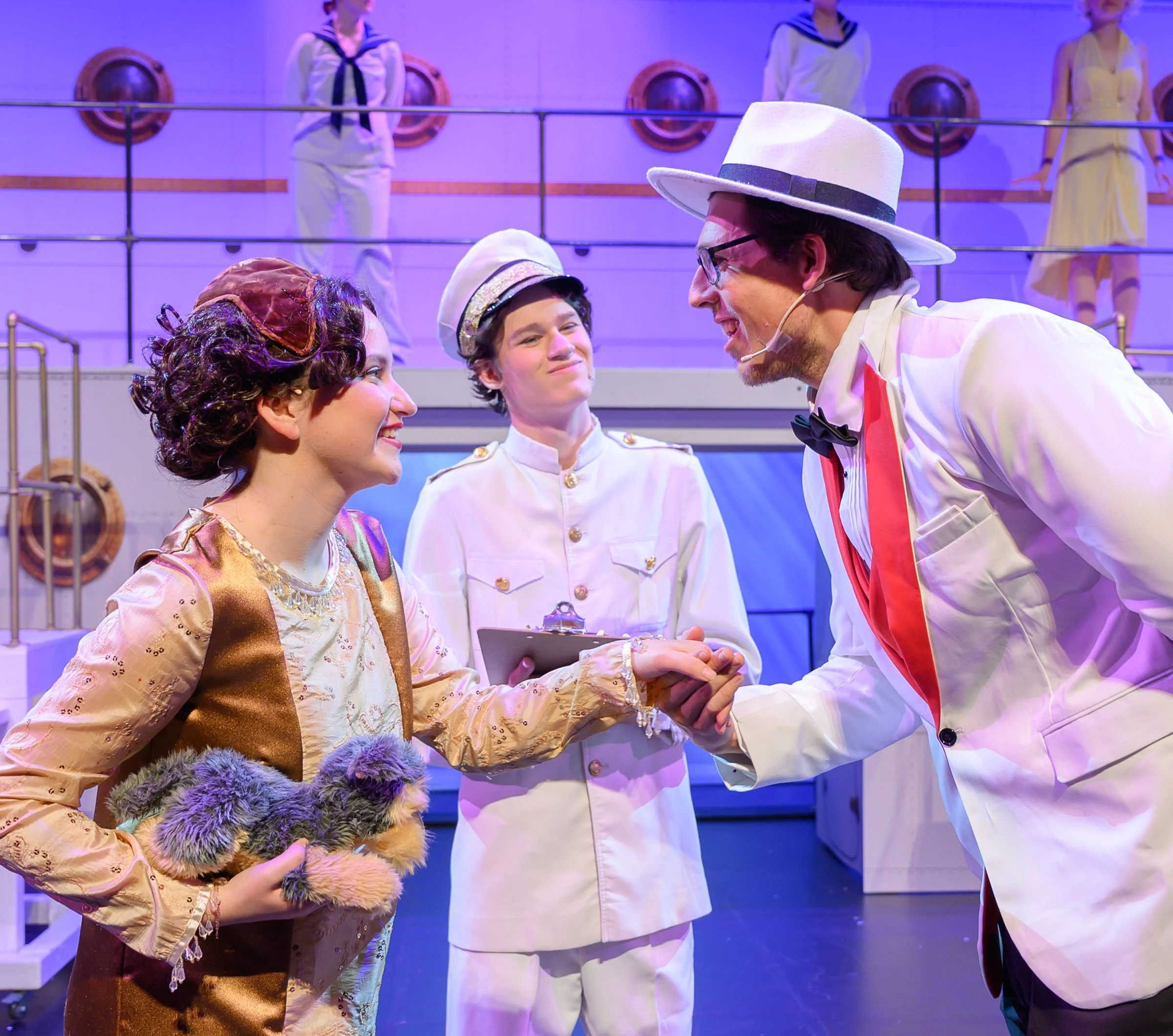 People in costume on stage, with women in white sailor outfits and a man in a white suit with a large white hat, black glasses, and red suspenders, engaging in a theatrical performance.
