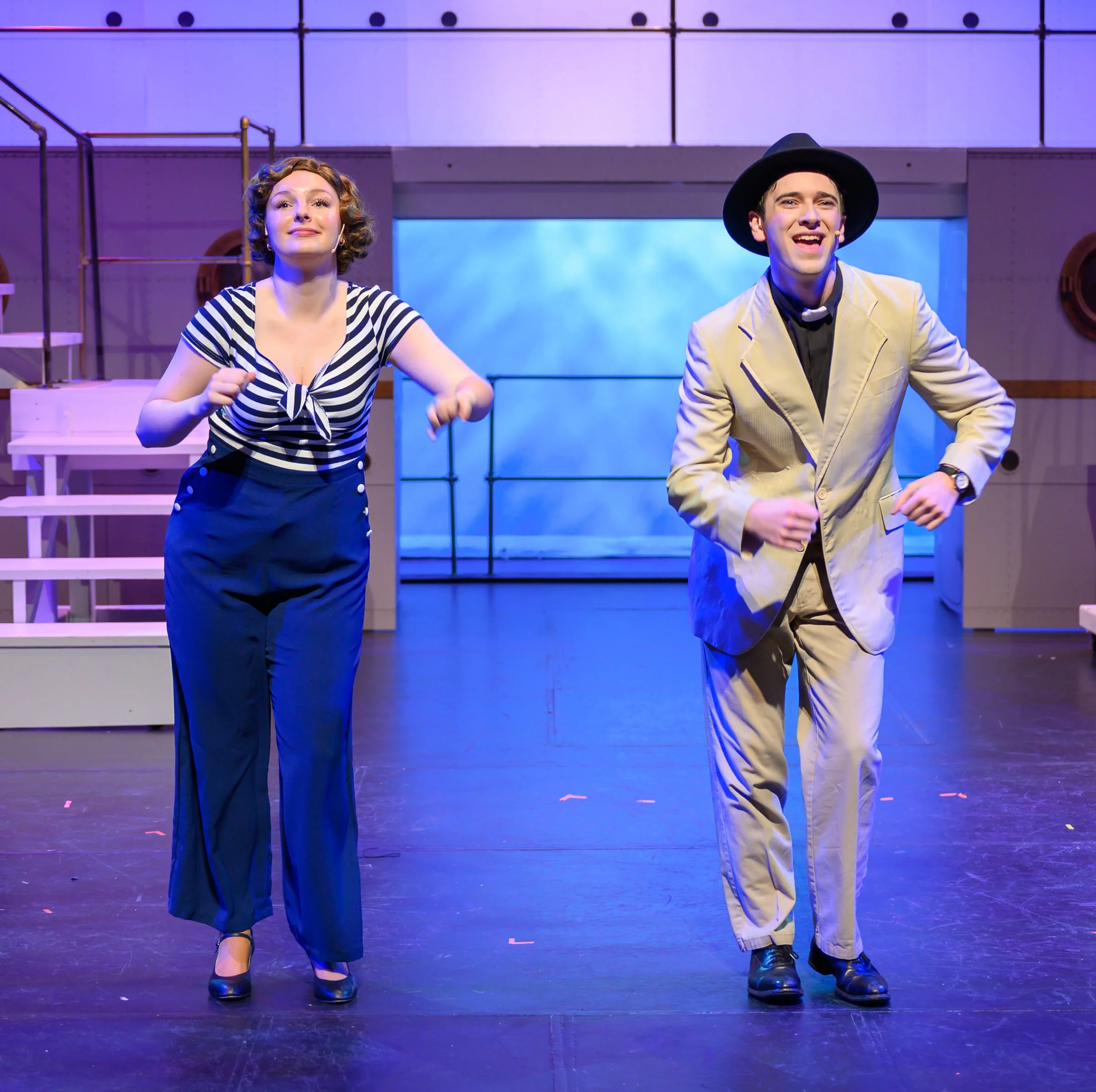 Two performers, a woman with curly hair in a striped shirt and dark pants, and a man with a black hat and beige suit, smiling and dancing on stage with purple and blue lighting.