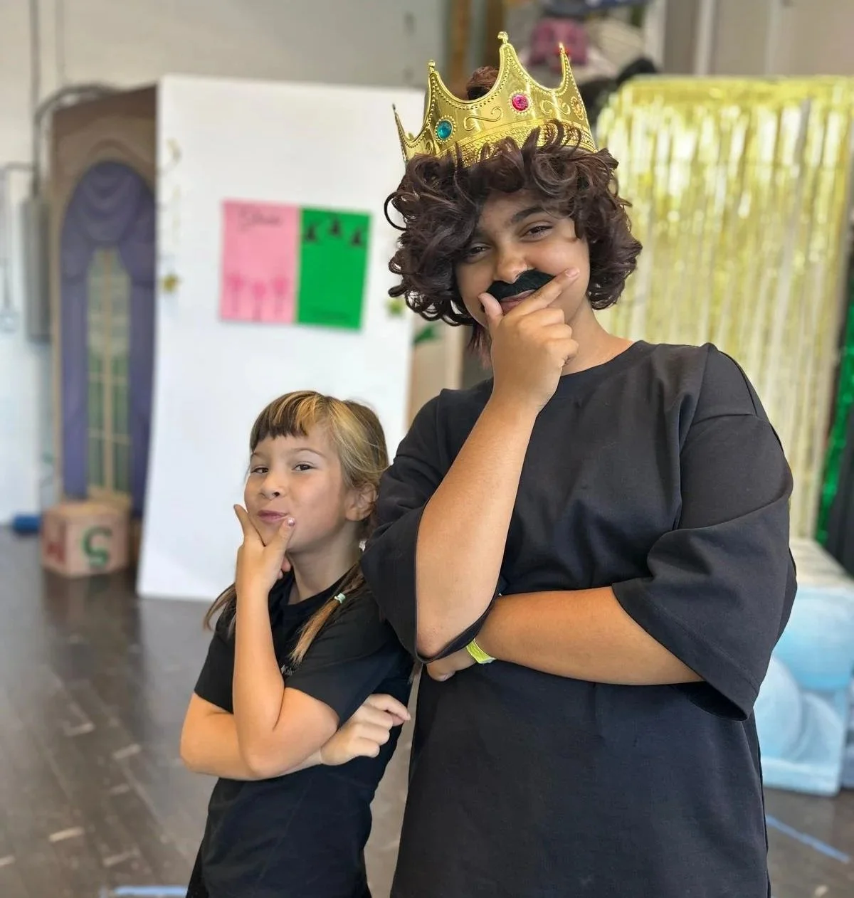 Two children dressed as royalty, one with a crown and mustache mask, striking playful poses indoors.