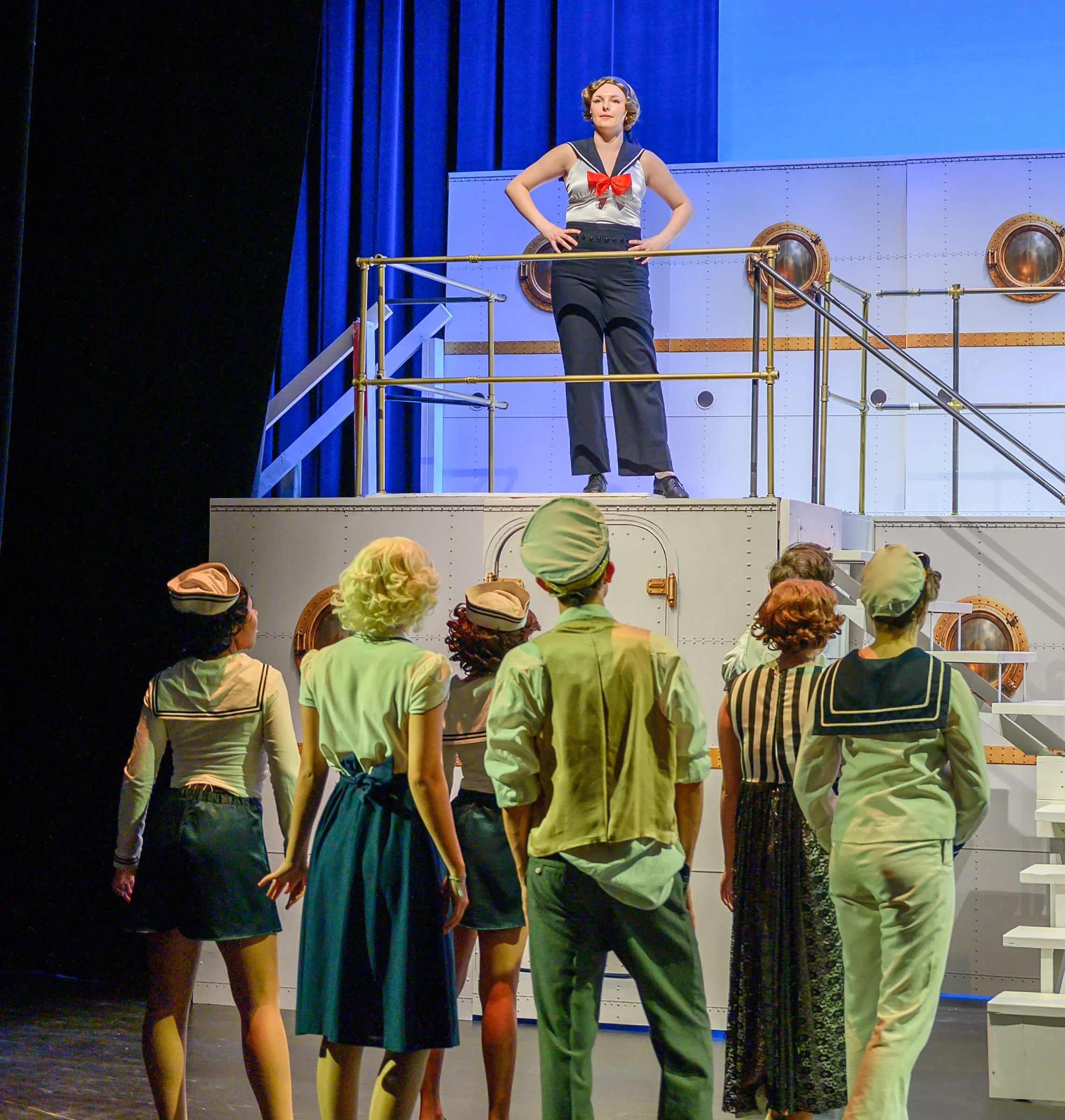 Women dressed in vintage swimwear standing on a stage with navy blue curtains and portholes, performing in front of an audience.