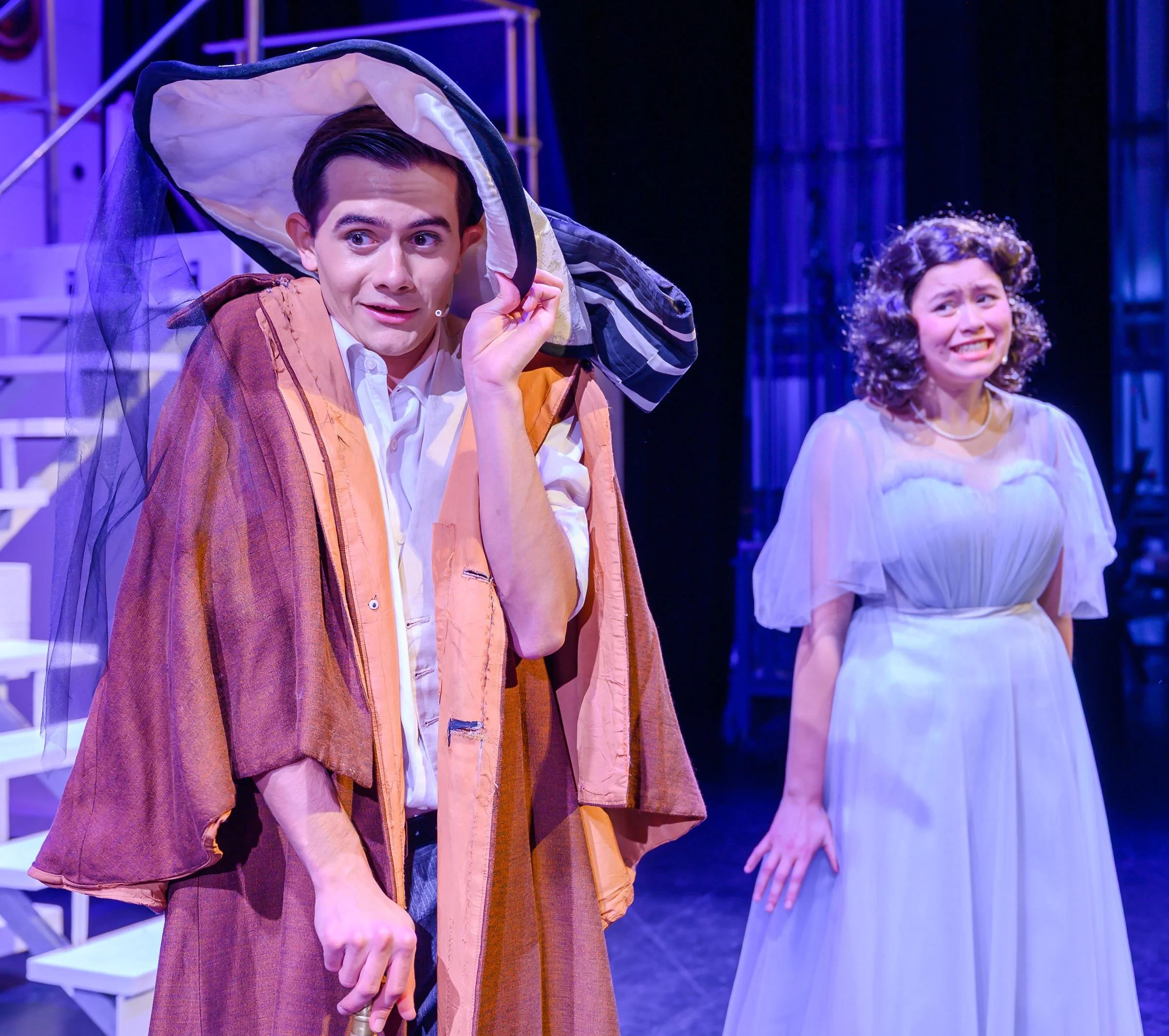 Two people performing on a stage with purple and blue lighting. The person on the left, a man with dark hair, is holding a large costume or prop over his head and has a surprised or curious expression. The person on the right, a woman with curly hair