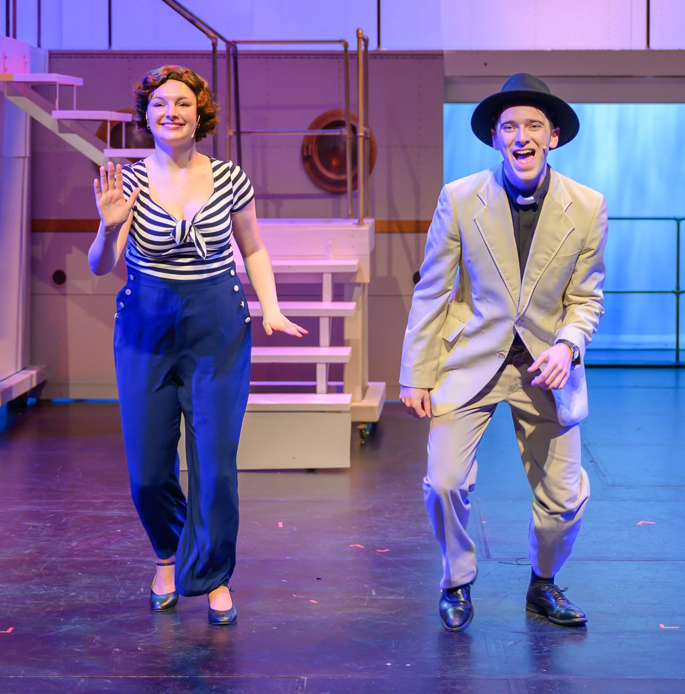 Performers on stage, a woman waving, dressed in a striped top and high-waisted pants, and a man in a beige suit and large hat, smiling and dancing.