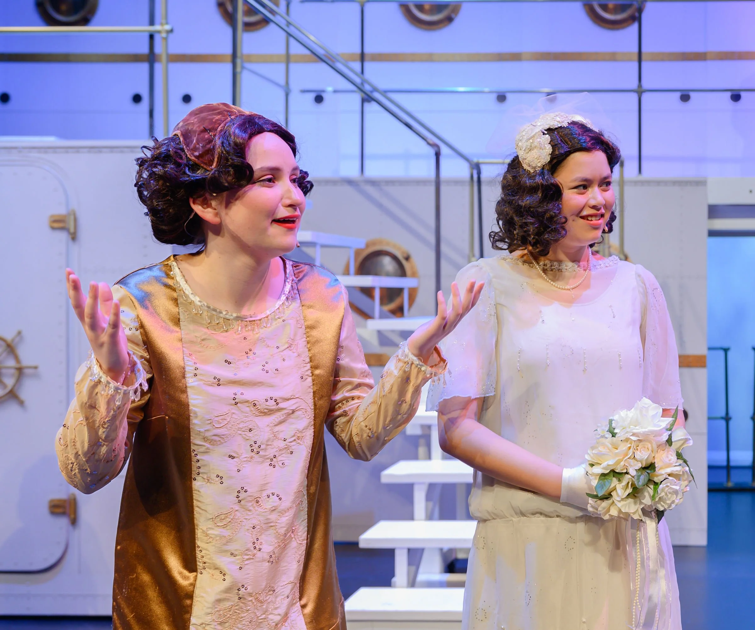 Two women dressed in vintage clothing, with curled hairstyles and matching headpieces. They are smiling and appear to be in a theatrical or costume setting with industrial-style background elements.