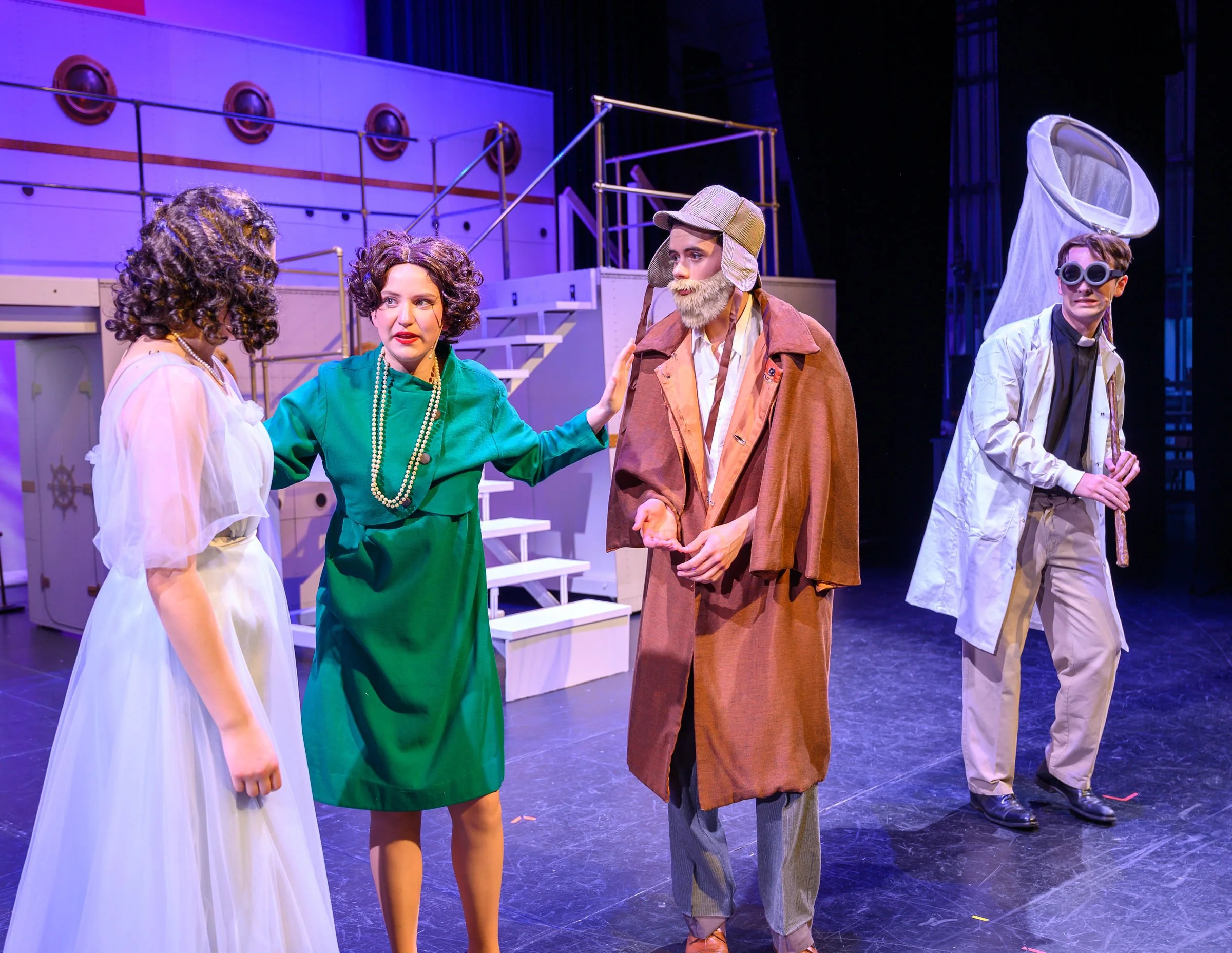Theatrical scene with four actors dressed in costumes, engaged in a conversation on stage. Background includes set pieces with nautical theme elements.