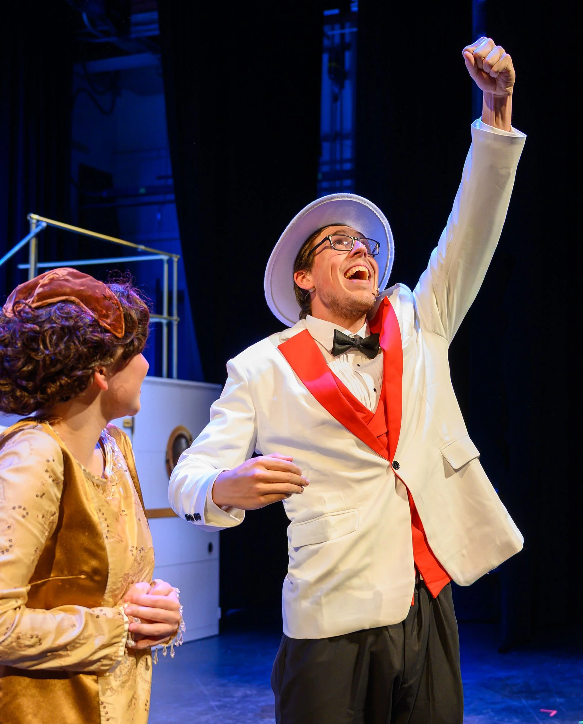 A man in a white tuxedo with red accents, black bow tie, glasses, and a white hat, smiling and raising his right fist in the air during a performance, with a woman in a gold dress and a mask in the foreground and a stage with equipment in the backgro