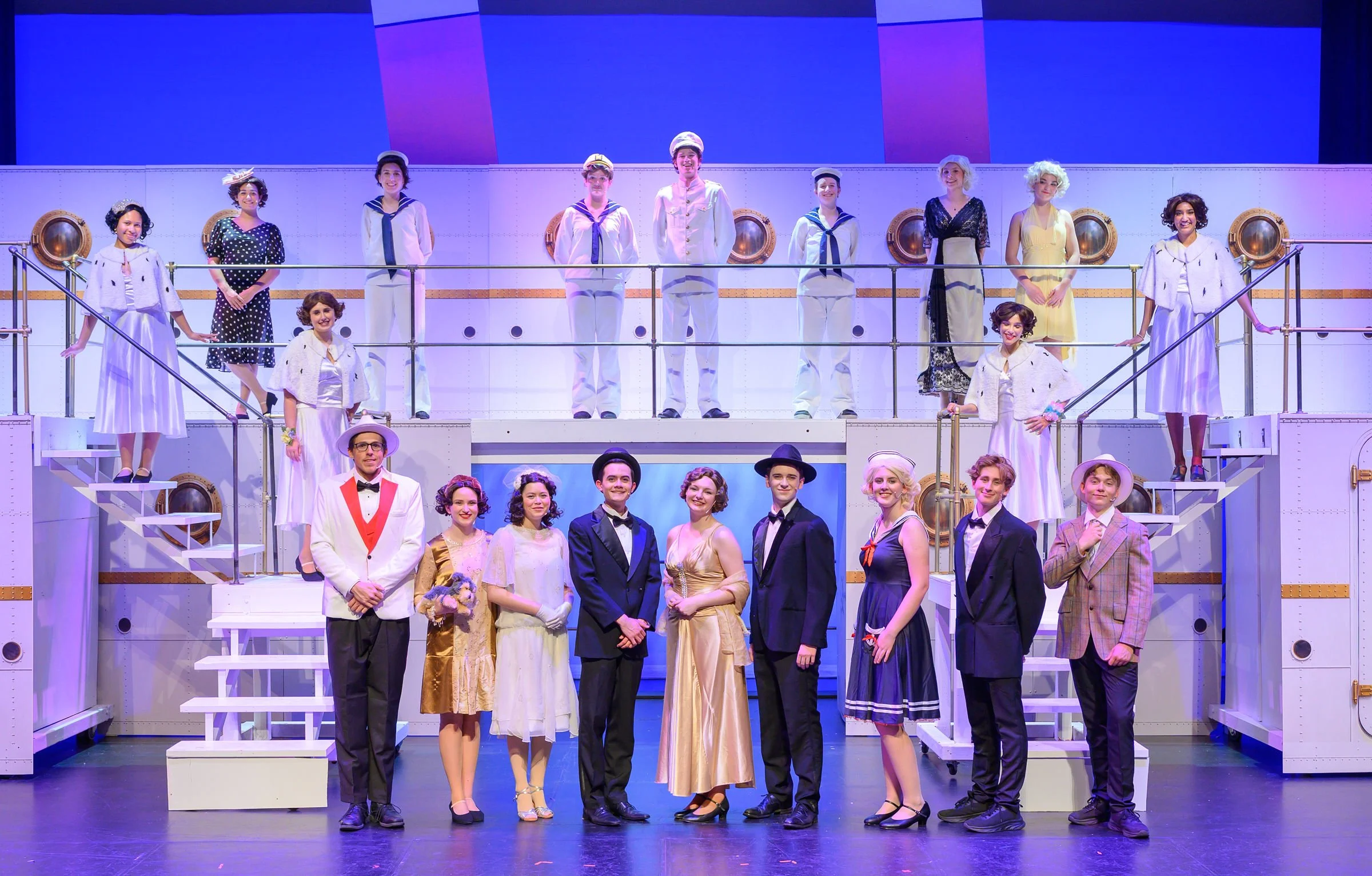 A group of people dressed in vintage costumes staged on a theater set resembling a ship's deck, with some standing on stairs and some on a lower level, in what appears to be a theatrical production.