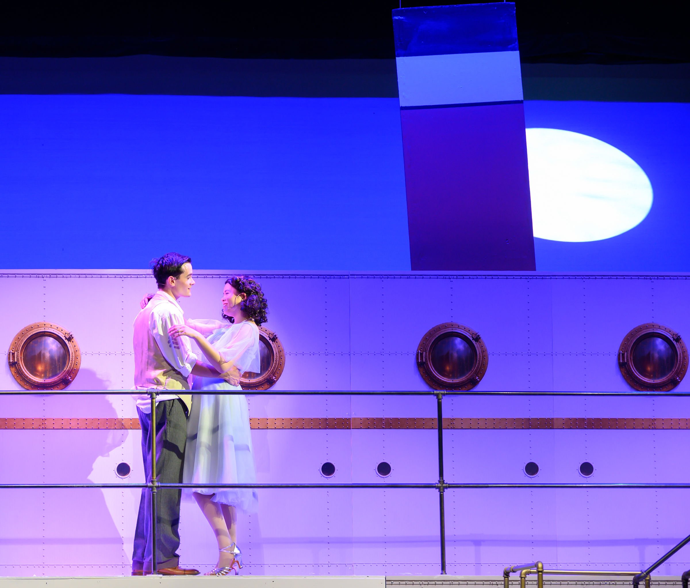 Two actors dressed in vintage clothing dancing on stage, with a blue background and round portholes in the set design.