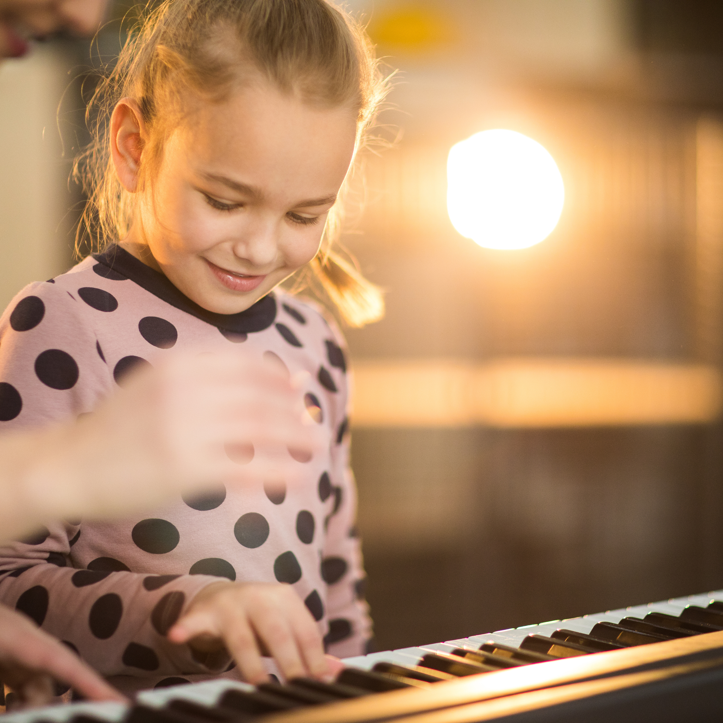 A young girl with a ponytail smiling as she plays the piano, with sunlight shining in the background.