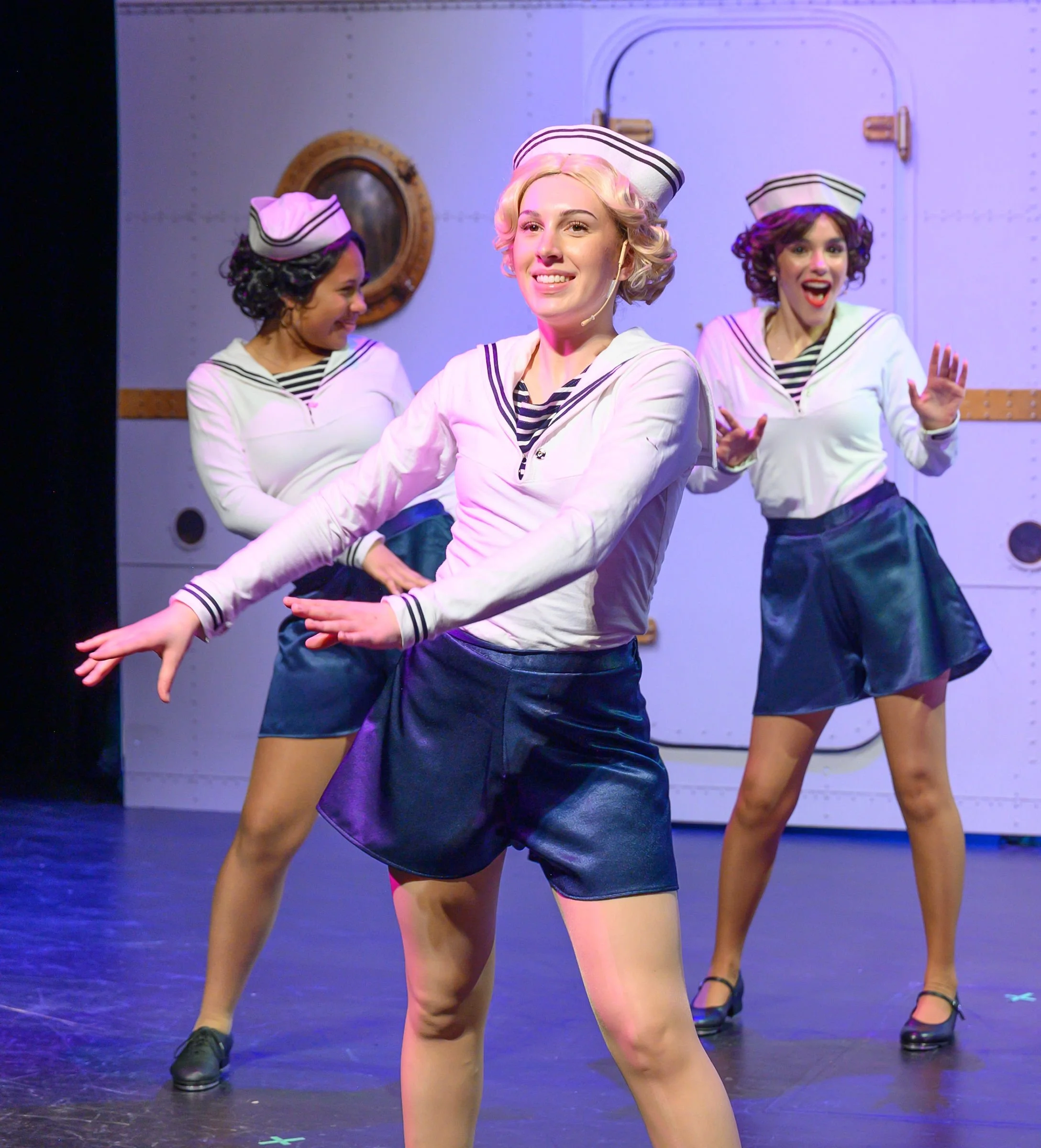 Three women dressed in sailor costumes performing a dance on stage. They are wearing sailor hats, striped shirts, and black skirts. The woman in the center is smiling, and the other two women are dancing in the background.