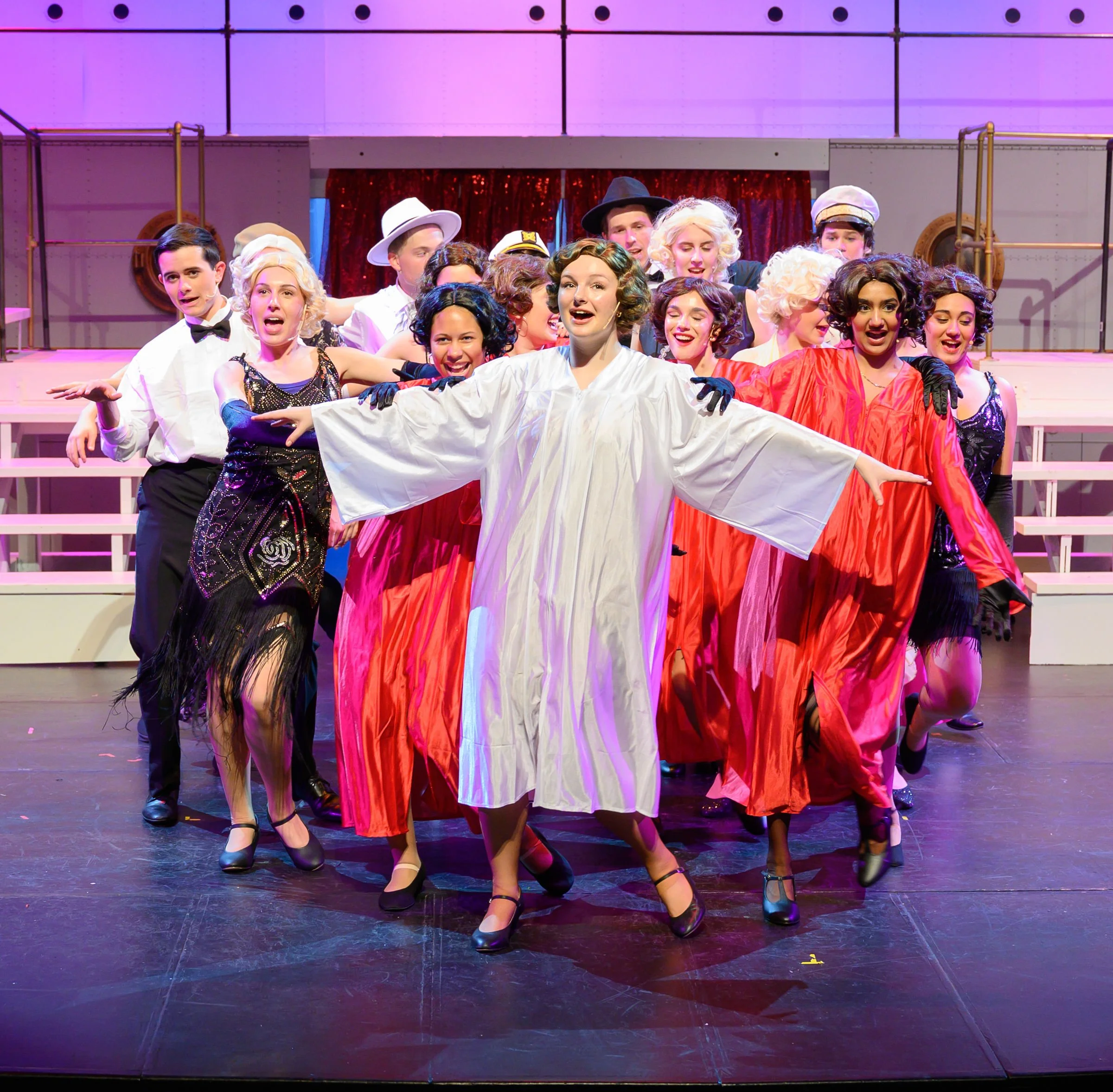 The image shows a group of diverse young actors performing on stage, dressed in vintage costumes reminiscent of the 1920s or 1930s, endearing and expressive. They are engaged in a theatrical scene, with some members singing or speaking and others pos
