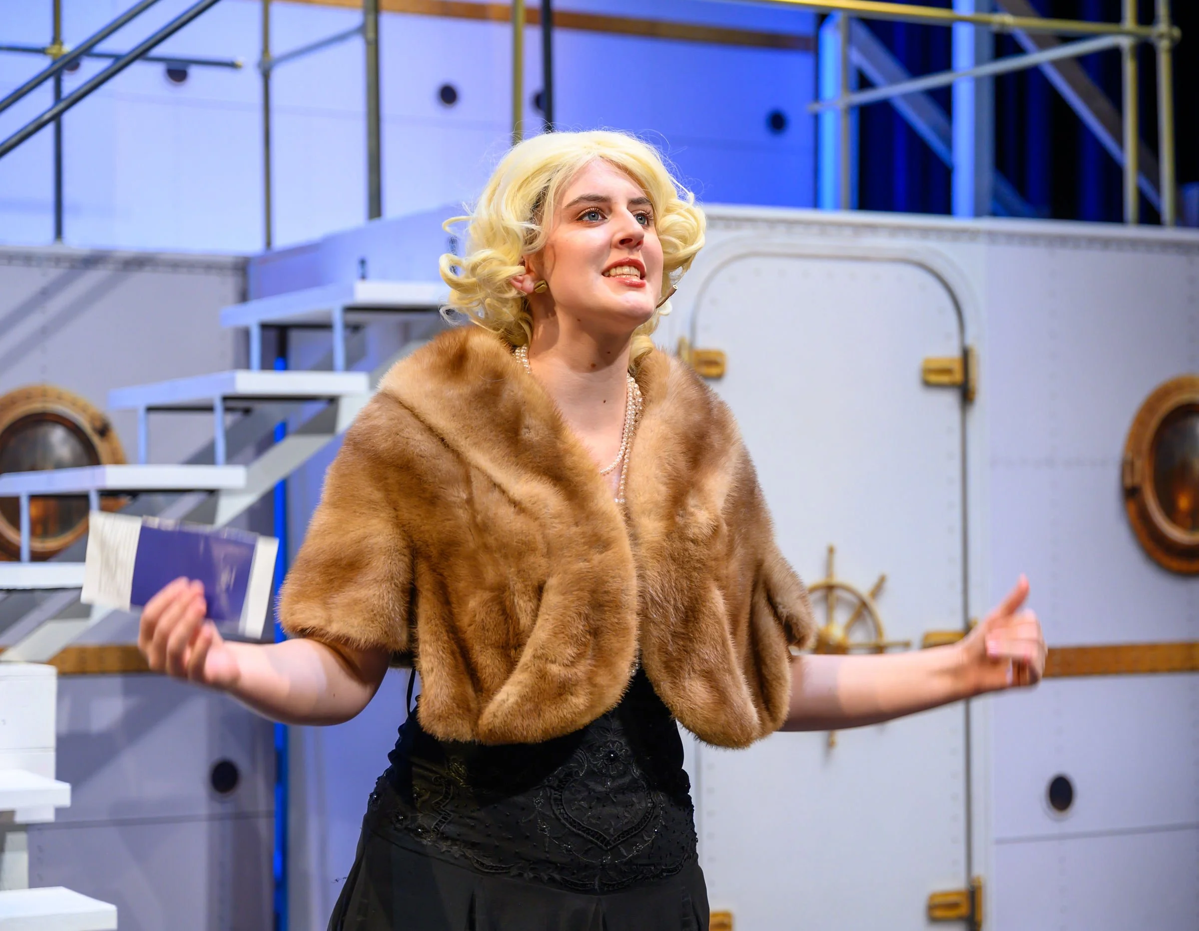 Woman with blonde curly hair wearing a fur coat and black dress, standing with arms outstretched in front of a stage set with white stairs and industrial-style metal structures.