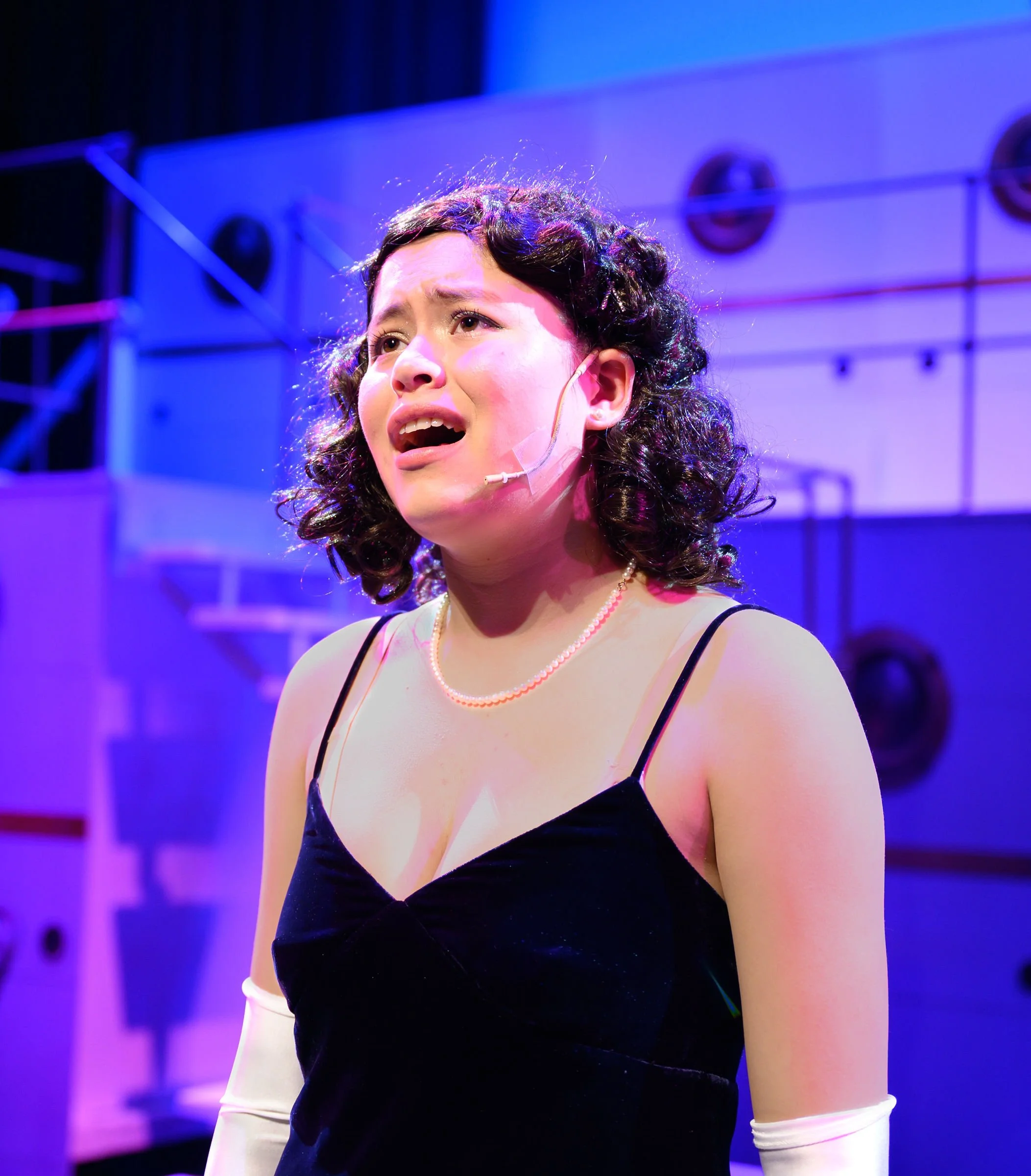 Close-up of a woman with curly hair and wearing a black dress with spaghetti straps, pearl necklace, white gloves, and a headset microphone, on stage with purple and blue lighting.