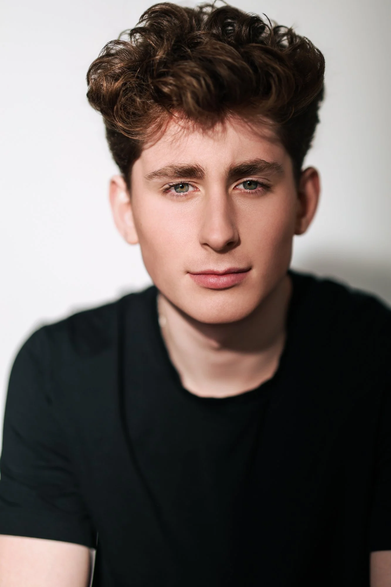 A young man with curly, reddish-brown hair and fair skin leaning against a brick wall, wearing a black shirt.