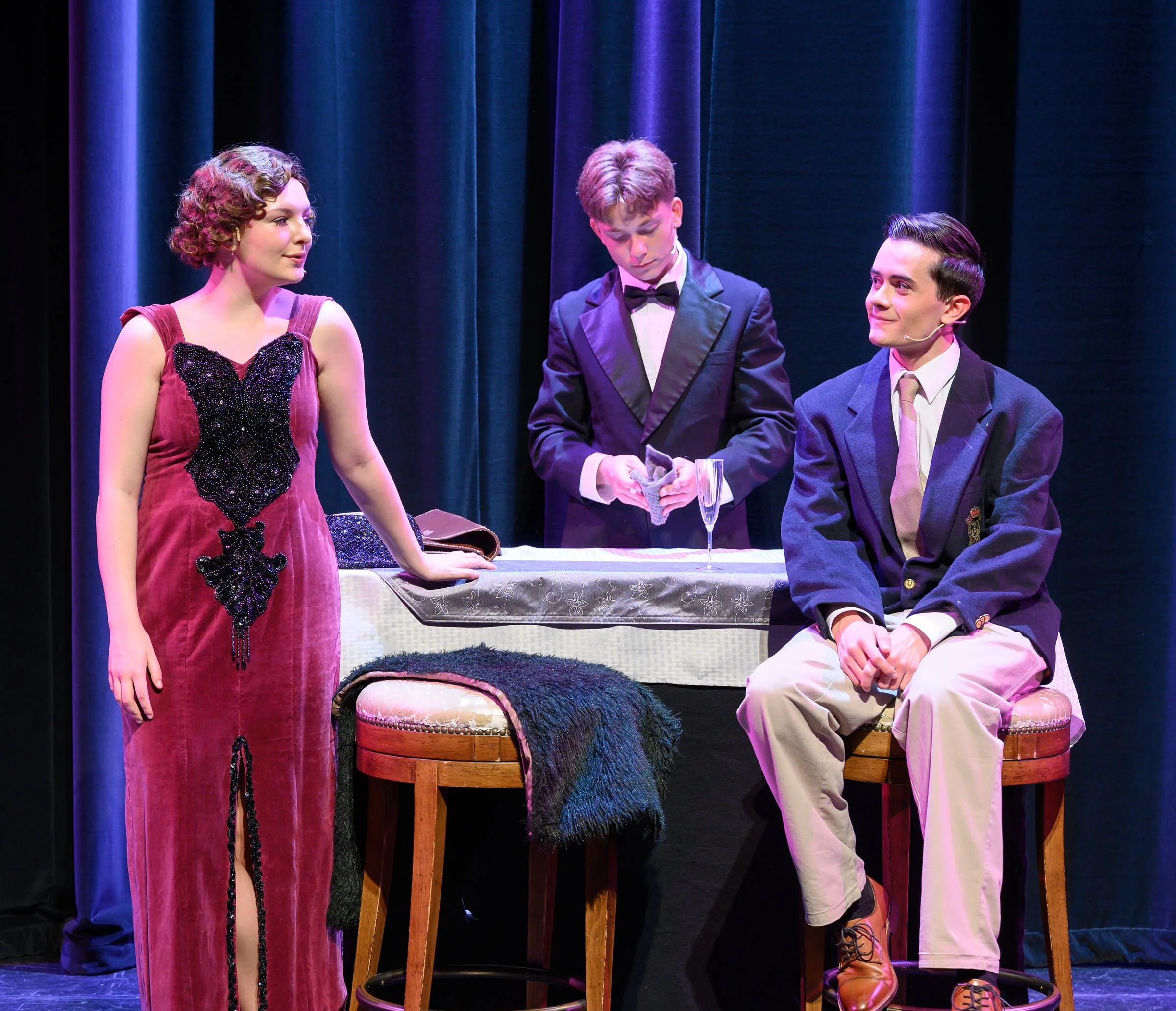 Three young actors in formal costumes on a stage with dark curtains in the background. One woman in a purple dress stands on the left, two men in tuxedos sit on a bench to her right, engaging in a performance.