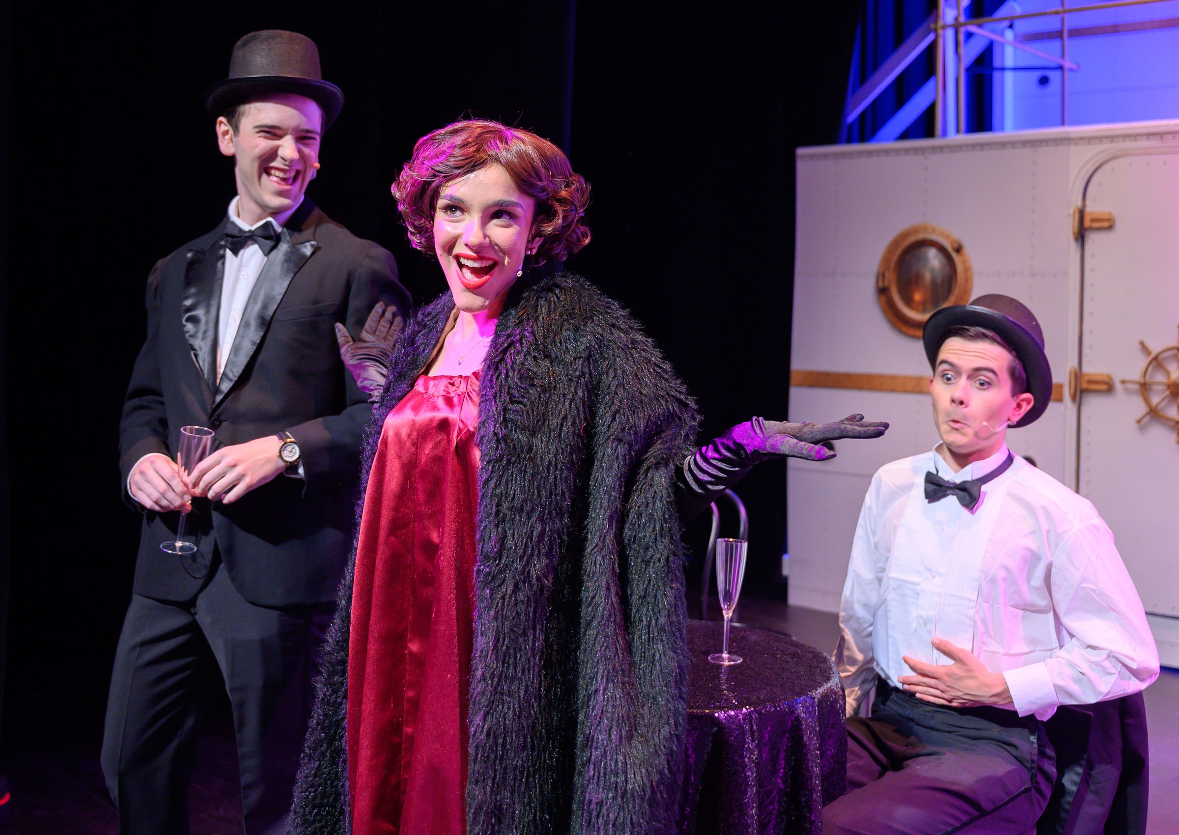 Three actors on stage in a theatrical performance, one man and two women, dressed in vintage costumes with humorous expressions.