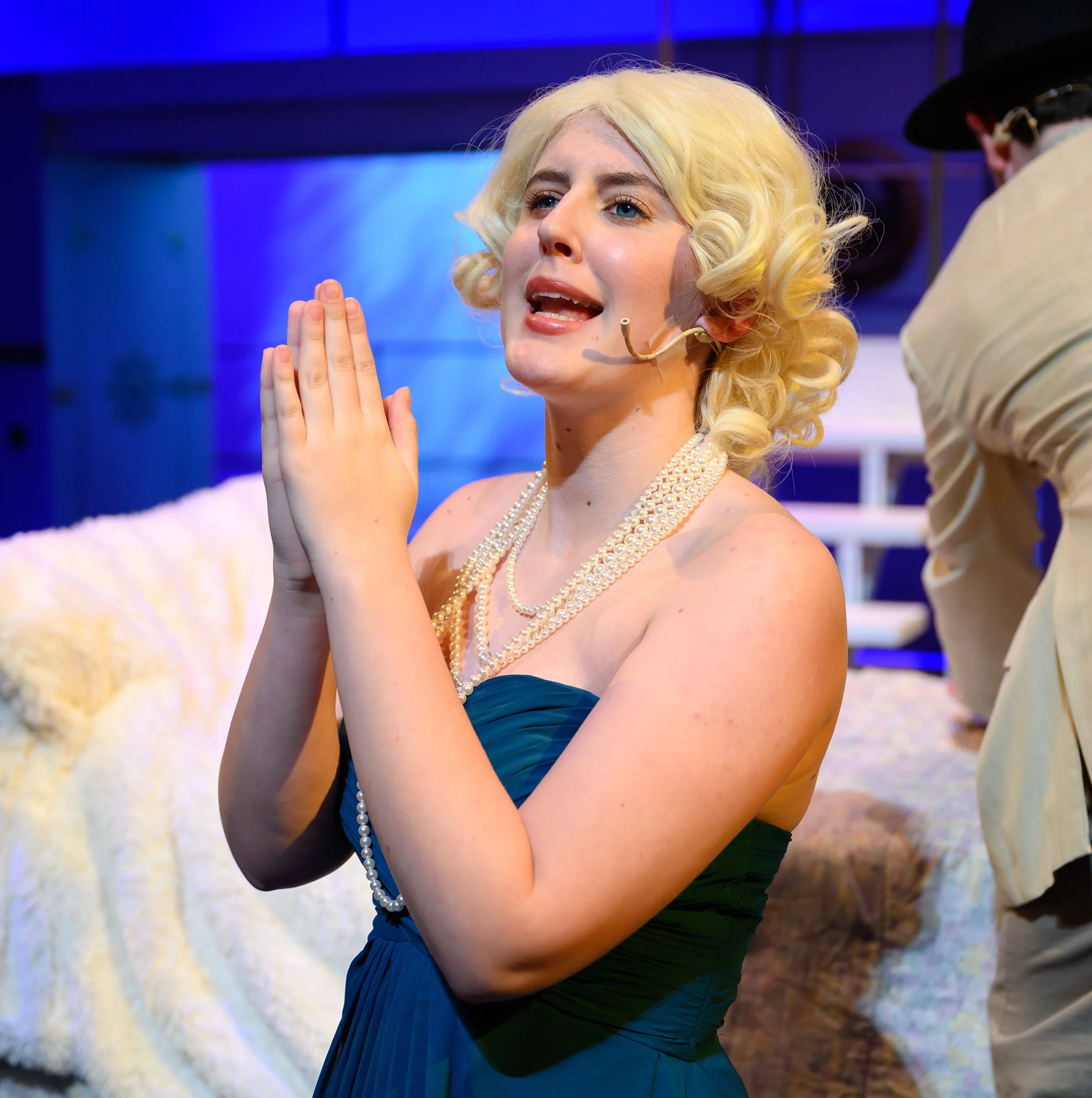 A woman with blonde, curly hair wearing a tuxedo dress, a pearl necklace, and a microphone headset, appears to be praying or speaking passionately with her hands pressed together. She is on stage with a blue and white abstract background.
