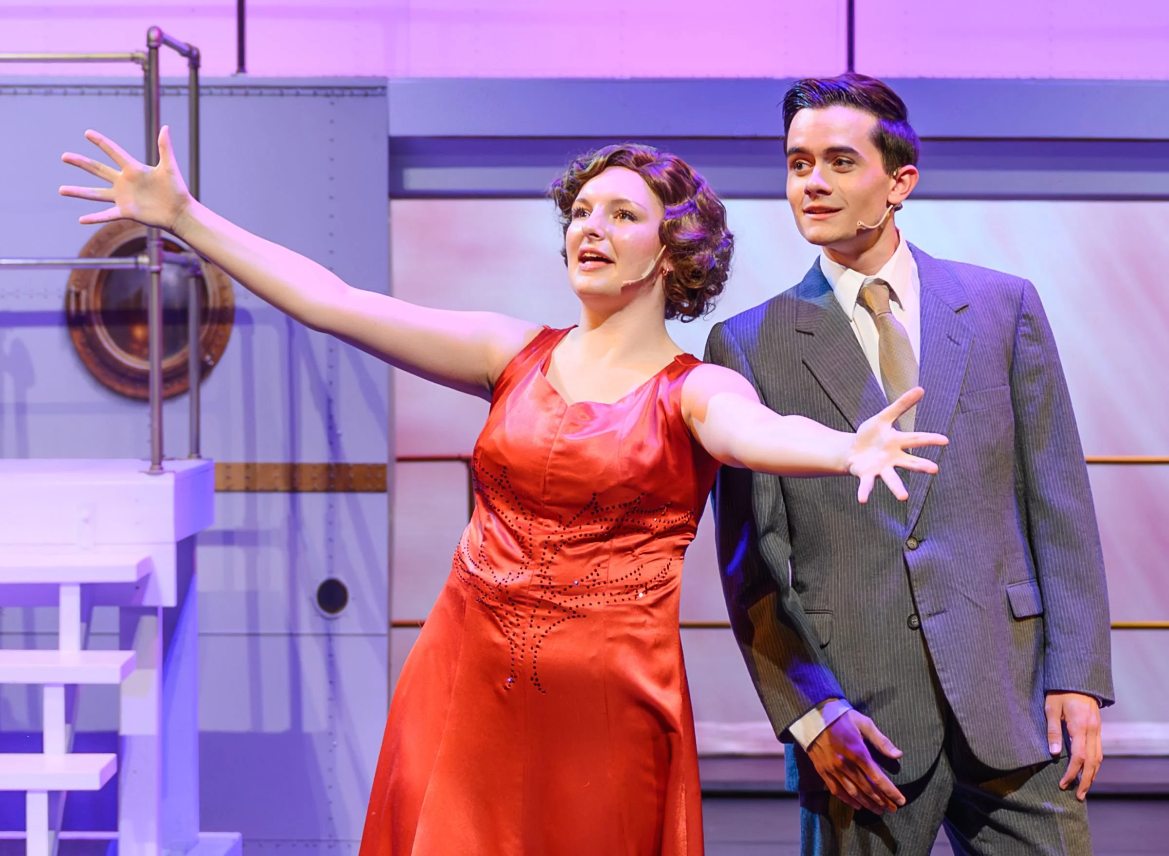 A woman in a red dress and a man in a gray suit with a microphone headset stand on a stage; the woman is gesturing outward with her arms, and the man is looking at her with a slight smile.