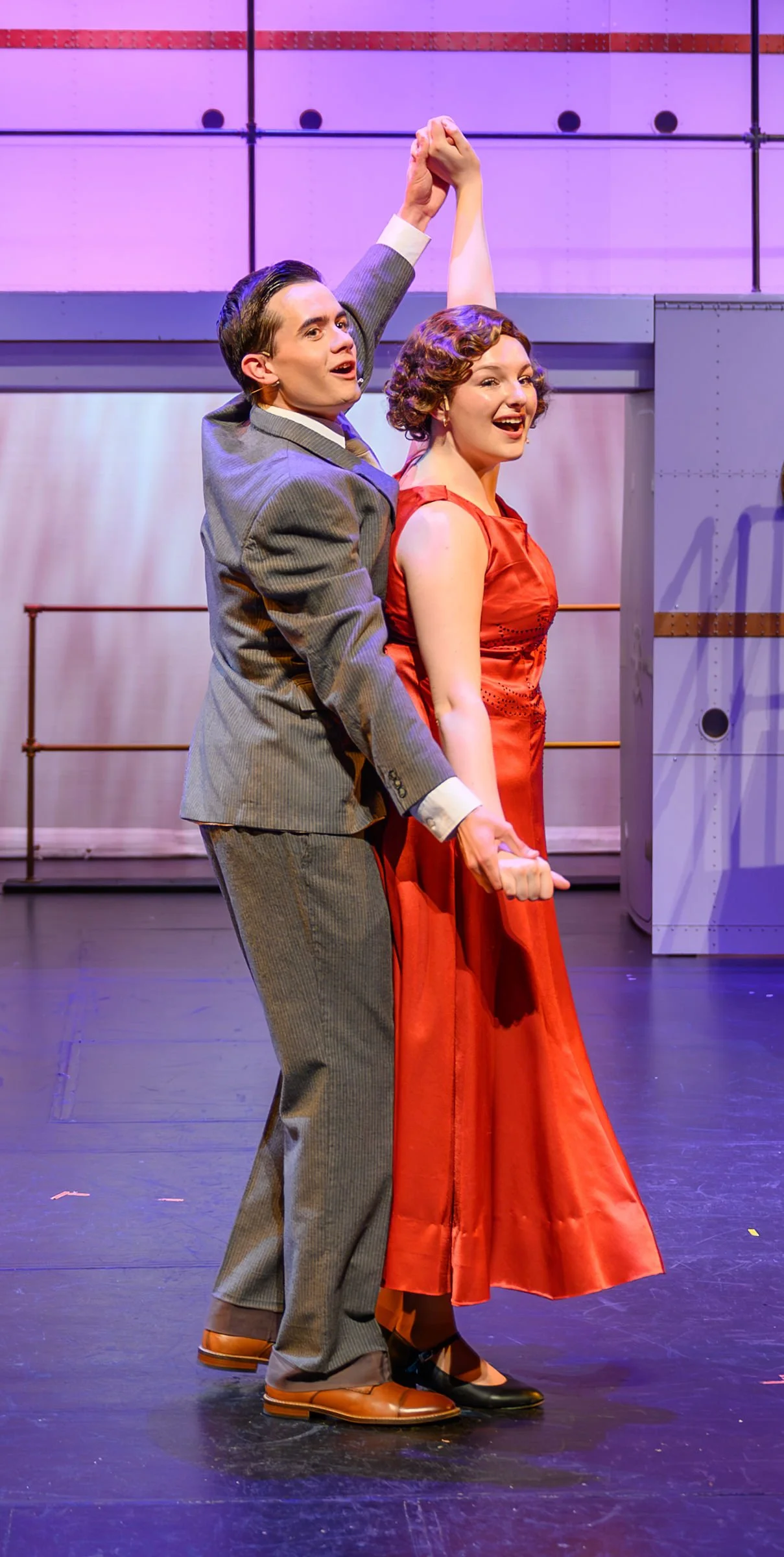 Two actors performing on stage, one is a man in a gray plaid suit and the other is a woman in a red dress, holding hands with their arms raised.