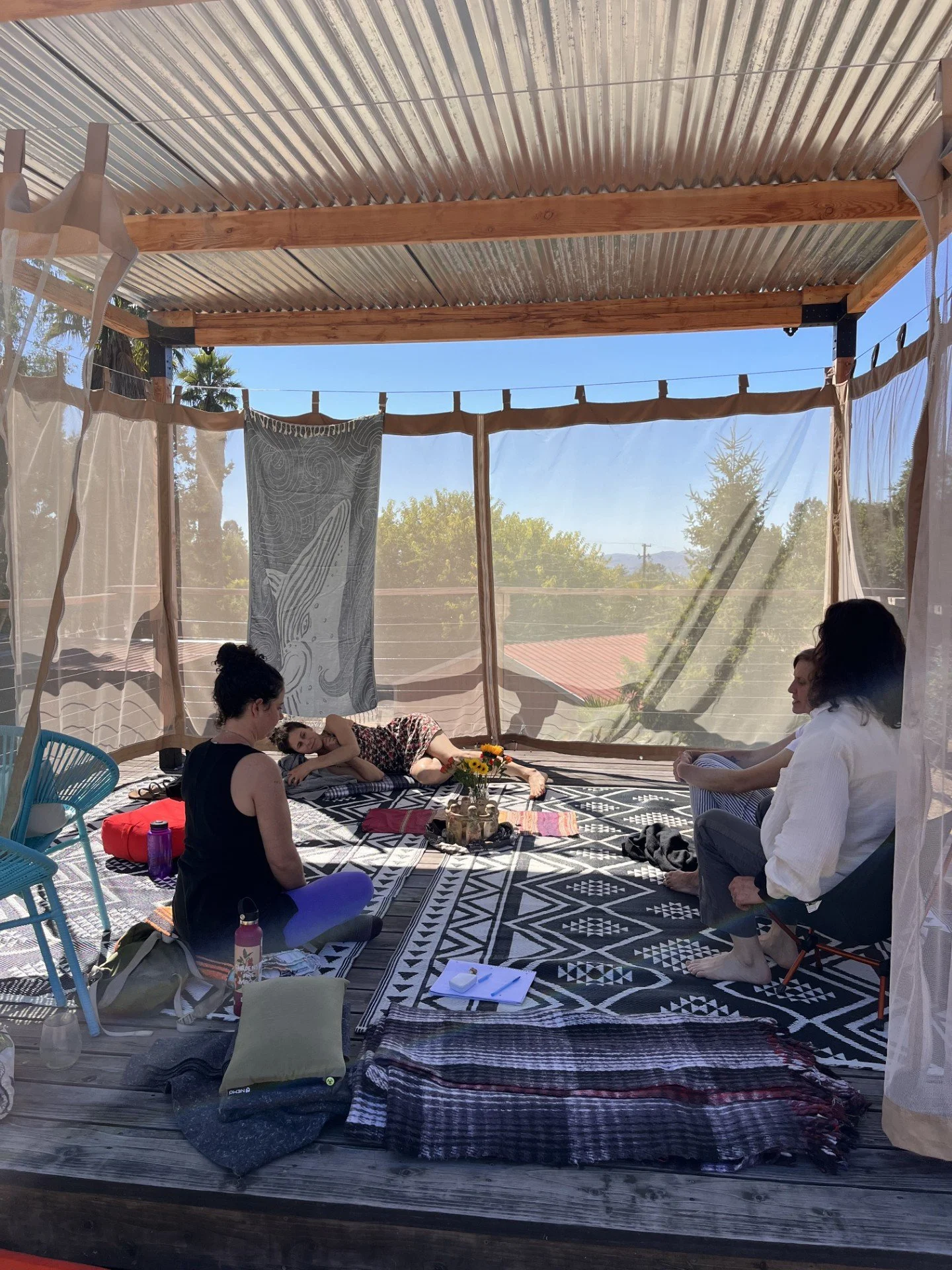 Group of women participating in a meditation or relaxing session in a covered outdoor space with a canopy, patterned rugs, and scenic view of trees and rooftops in the background.