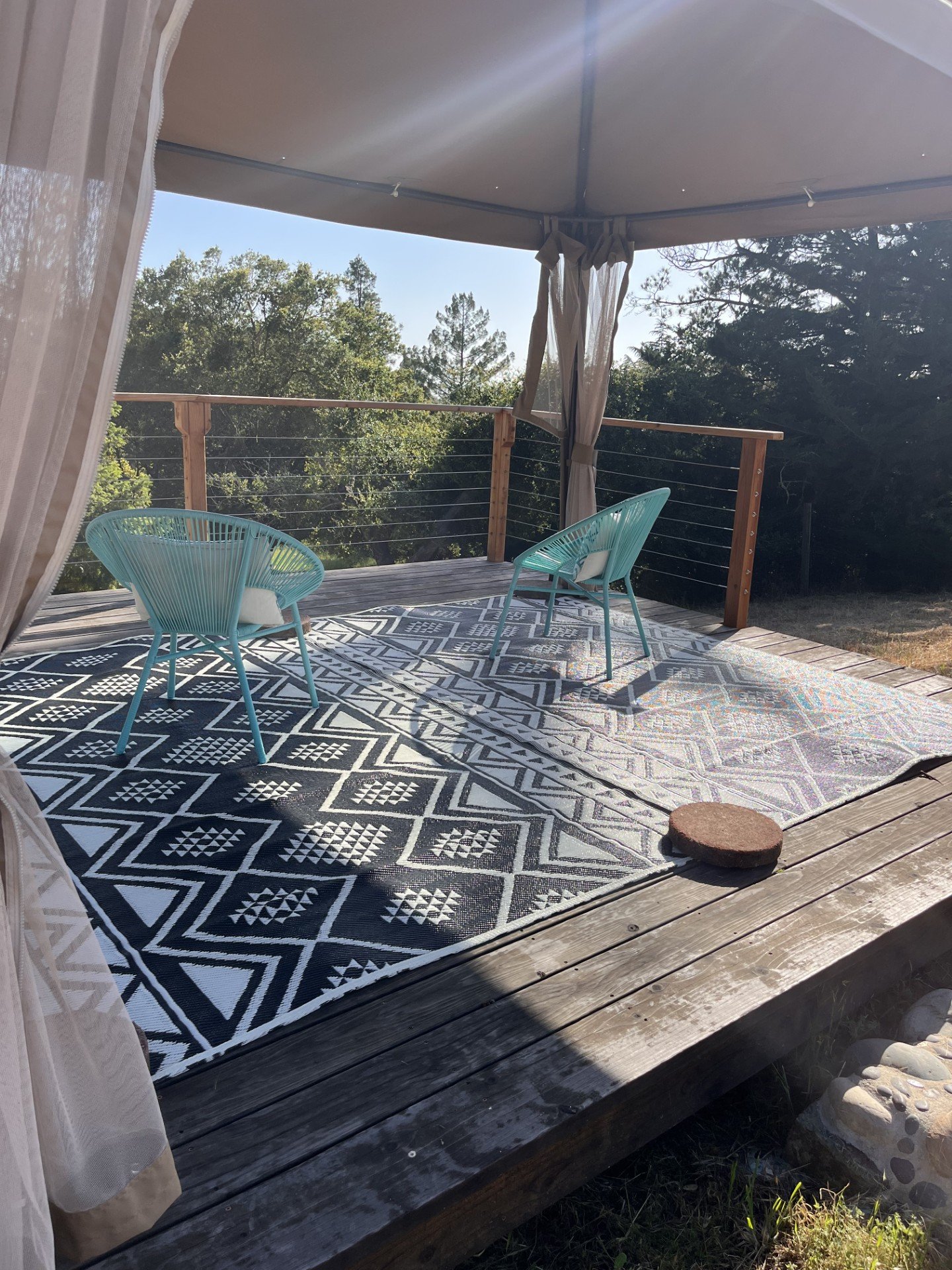 A screened outdoor porch with two teal chairs, a patterned outdoor rug, surrounded by trees and blue sky.