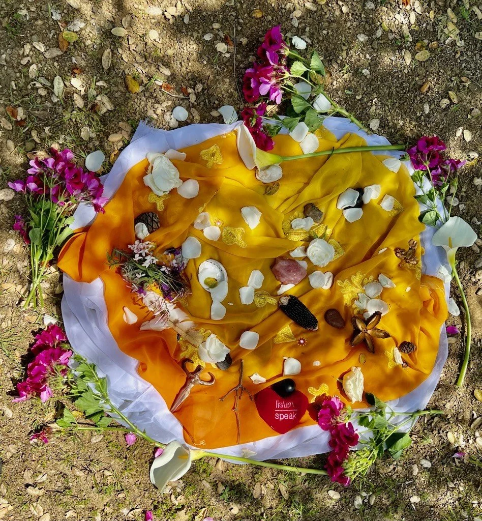 A memorial arrangement on the ground with colorful flowers, stones, candles, and fabric. The fabric is yellow, orange, and white, and there are pink and white flowers surrounding it. A small red heart-shaped object with the words "listen speak" is in