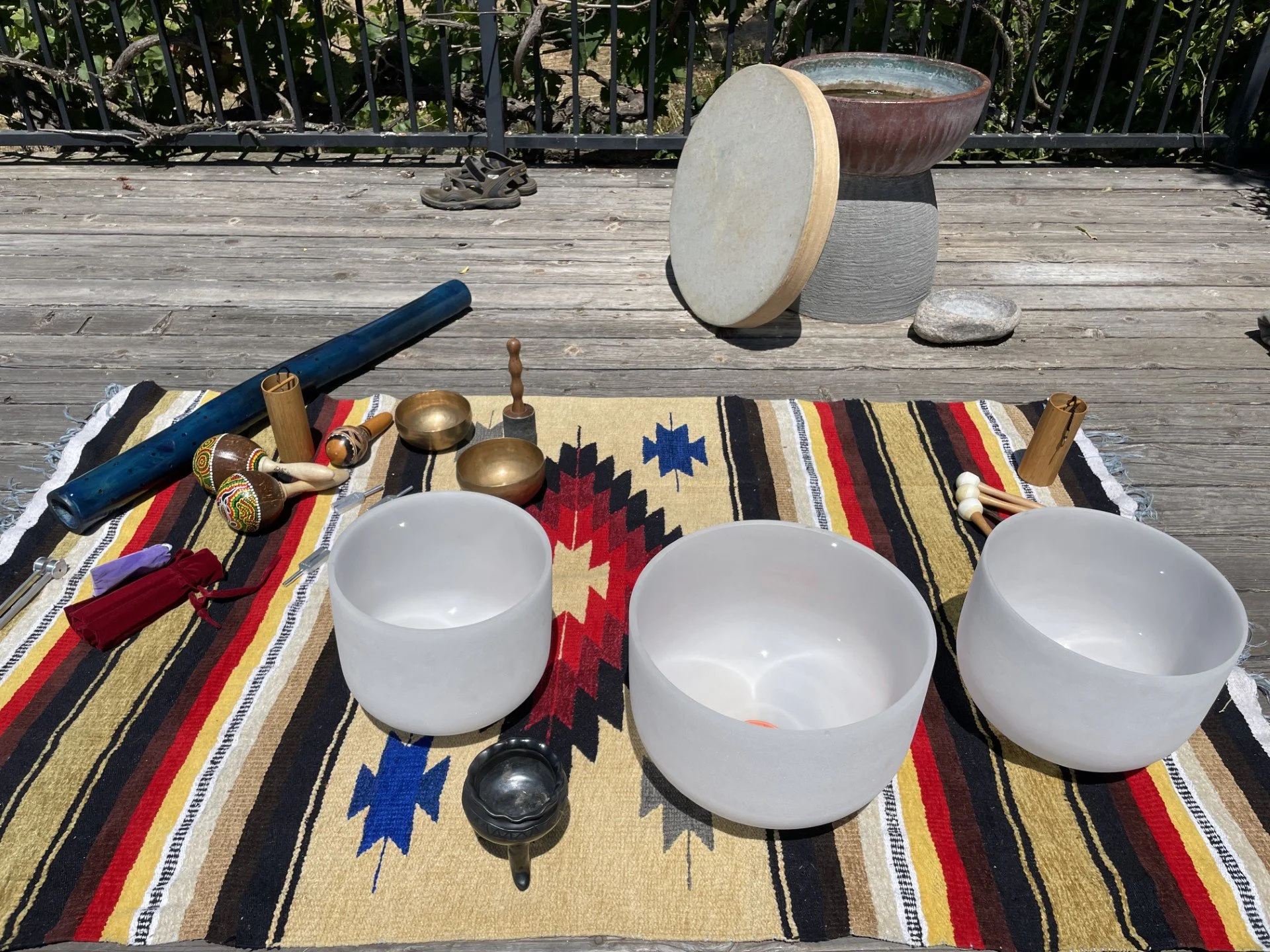 Musical instruments for a Native American or spiritual ceremony are laid out on a decorated cloth on a wooden deck outdoors. Items include three white singing bowls, a large drum, wooden percussion sticks, a tongue drum, a flute, and various small pe