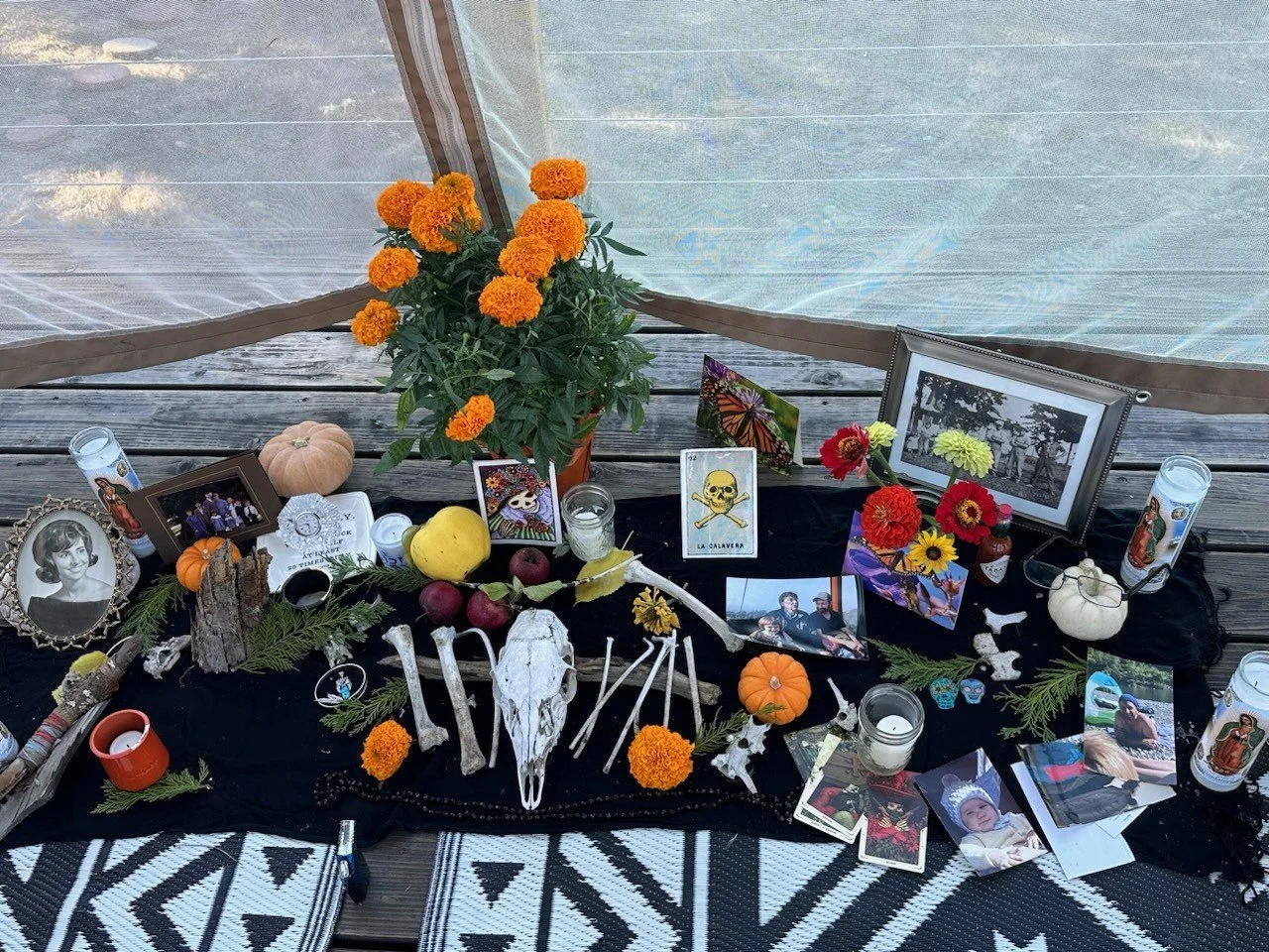 Day of the Dead memorial altar with flowers, candles, photographs, and decorative items on black fabric, placed on a wooden floor inside a tent.