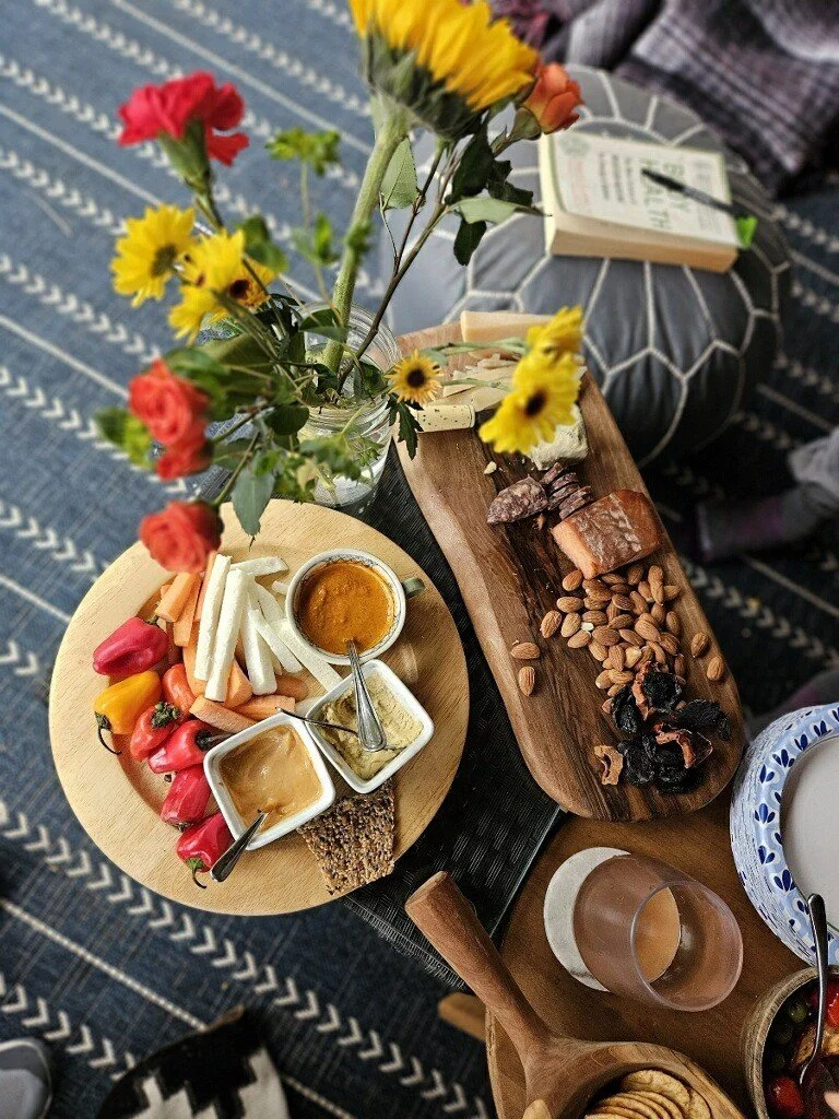 A table with a bouquet of colorful flowers, assorted cheeses, nuts, dried fruits, fresh vegetables, dips, and drinks is set on a patterned blue and white rug.