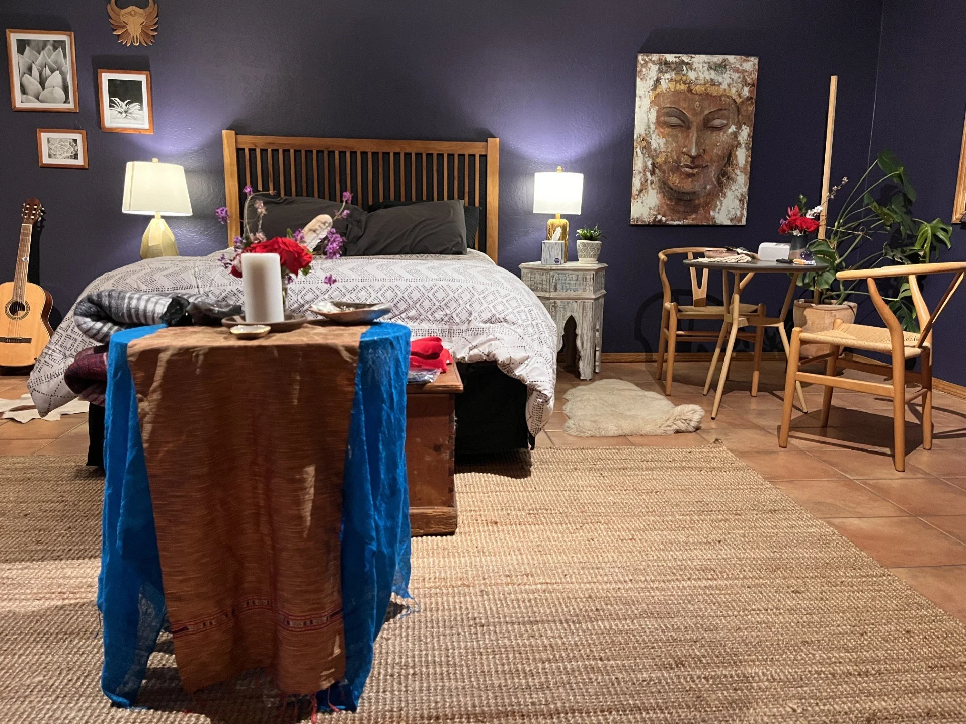 A cozy bedroom with a wooden bed frame, dark gray bedding, and two lamps on white nightstands. There is a guitar on the left, hanging art on the blue wall, a table with a floral arrangement, candle, and tray. To the right, a small round table with ch