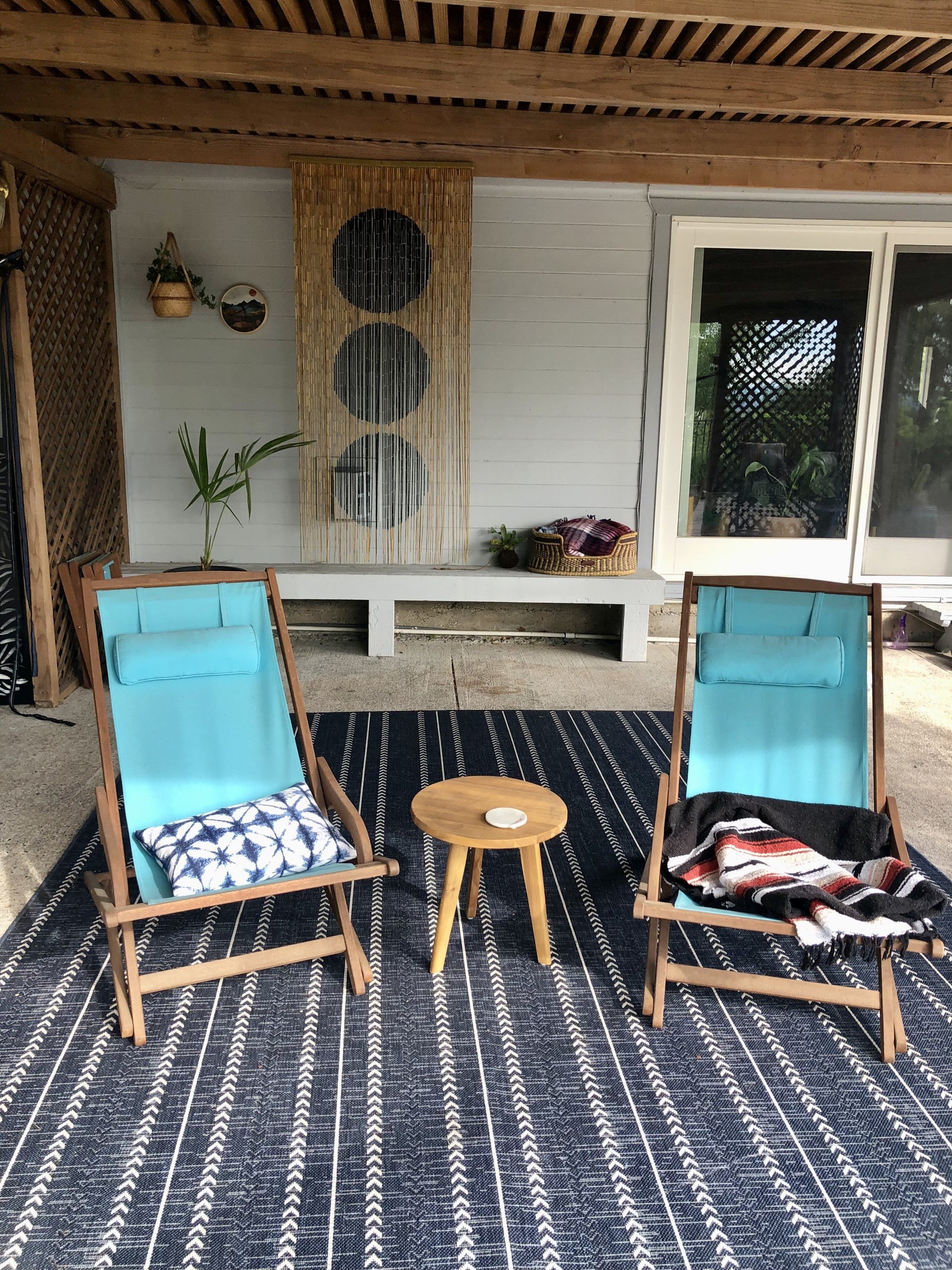 A cozy outdoor seating area with two wooden lounge chairs with turquoise cushions, a small round wooden table between them, a black and white patterned outdoor rug, a white wall with a sliding glass door, a large abstract wall hanging with three blac