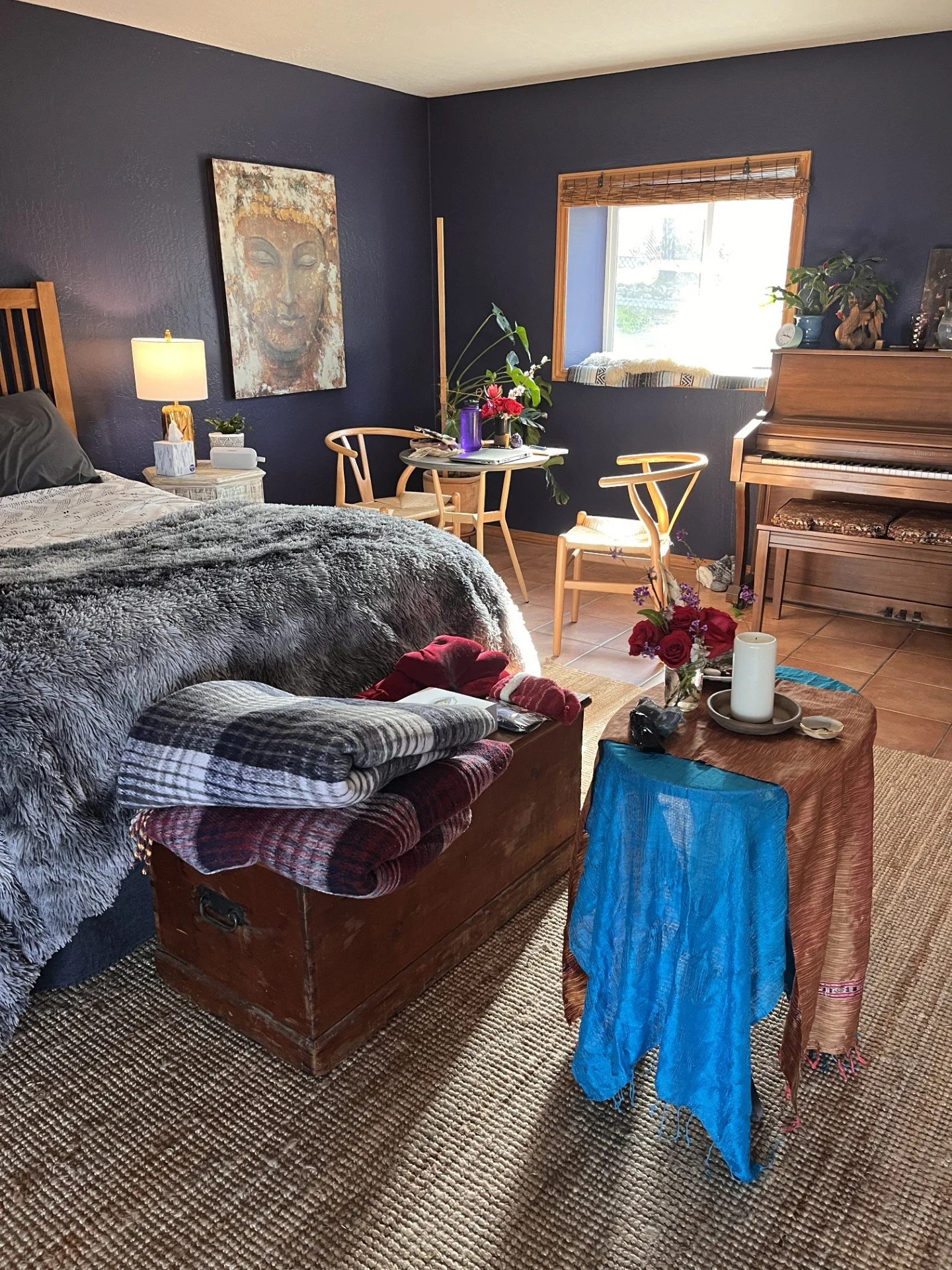 Modern bedroom with a dark blue wall, a bed with gray bedding, a wooden chest with folded blankets, a small table with chairs, a window, sacred decorative items, plant decorations, and a small table with candles and flowers.