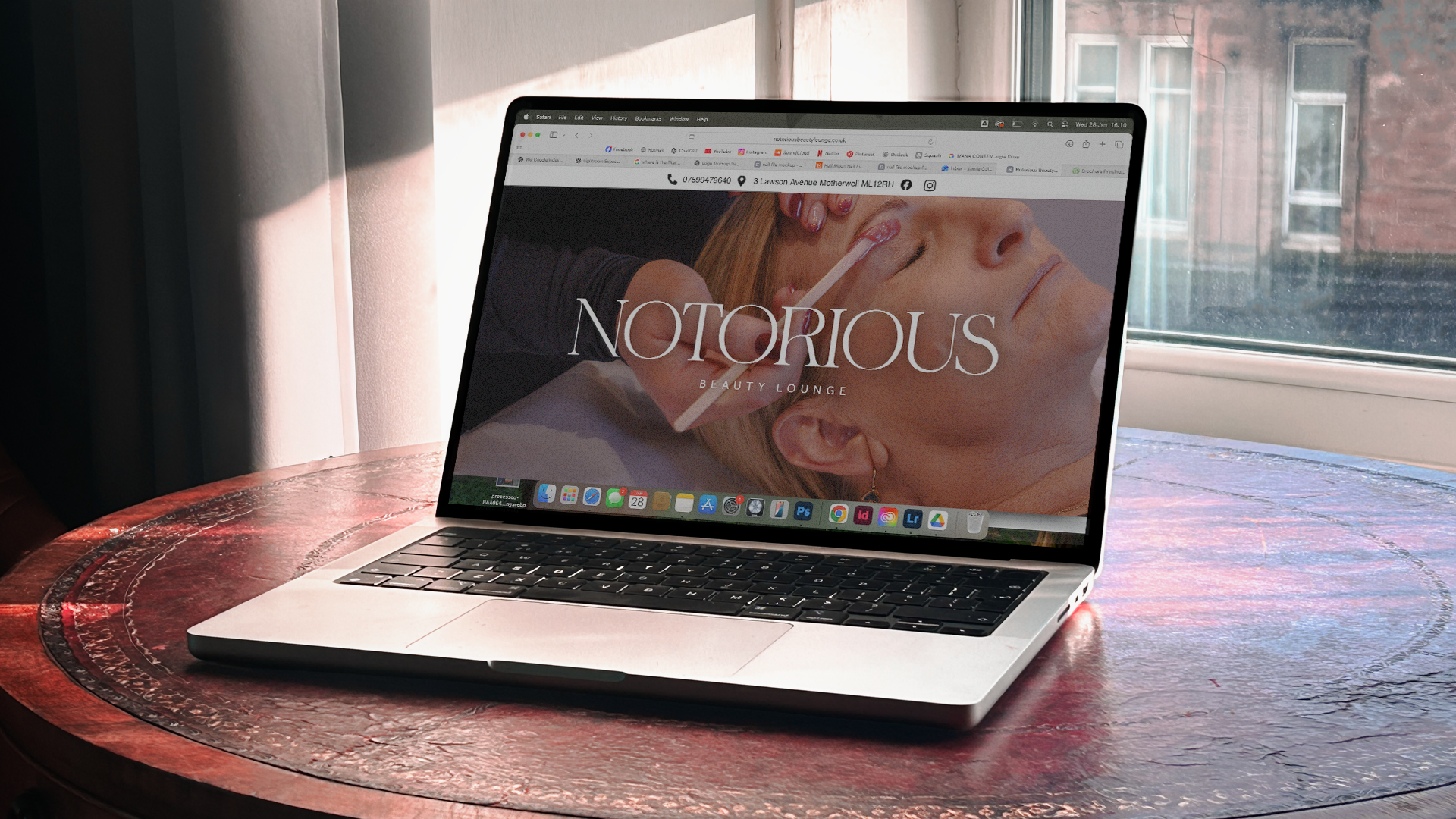 A MacBook laptop on a round wooden table near a window, displaying a website for Notorious Beauty Lounge with an image of a woman receiving a facial treatment.