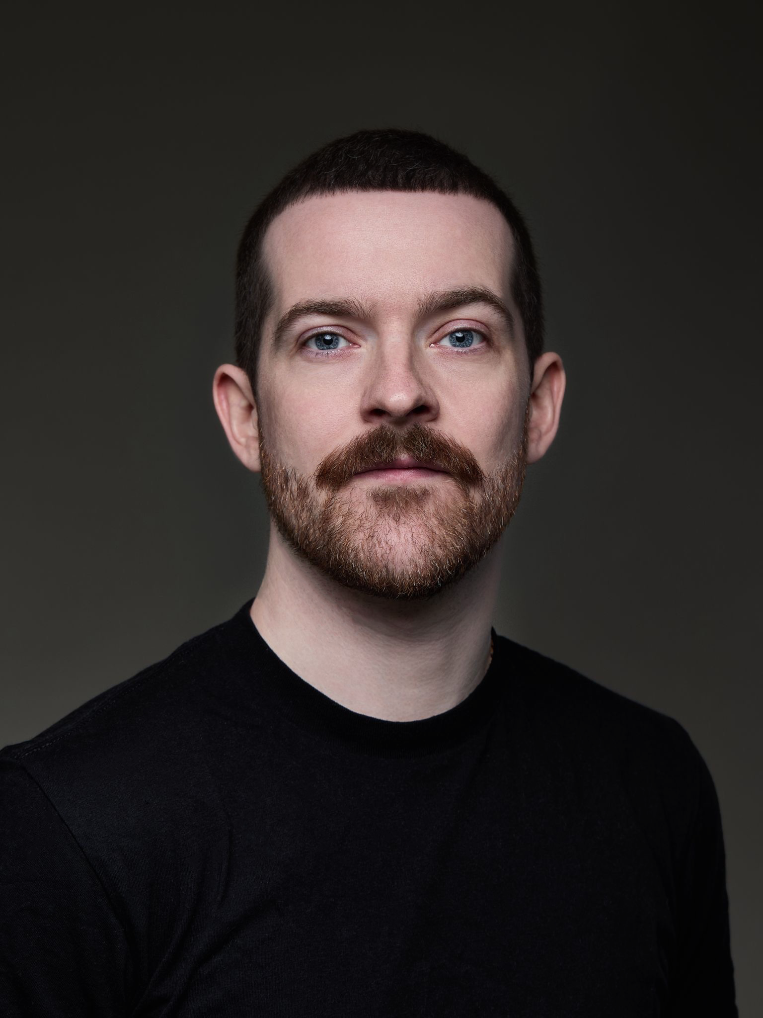 A portrait of a man with short dark hair, a beard, and blue eyes, wearing a black shirt, against a dark background.