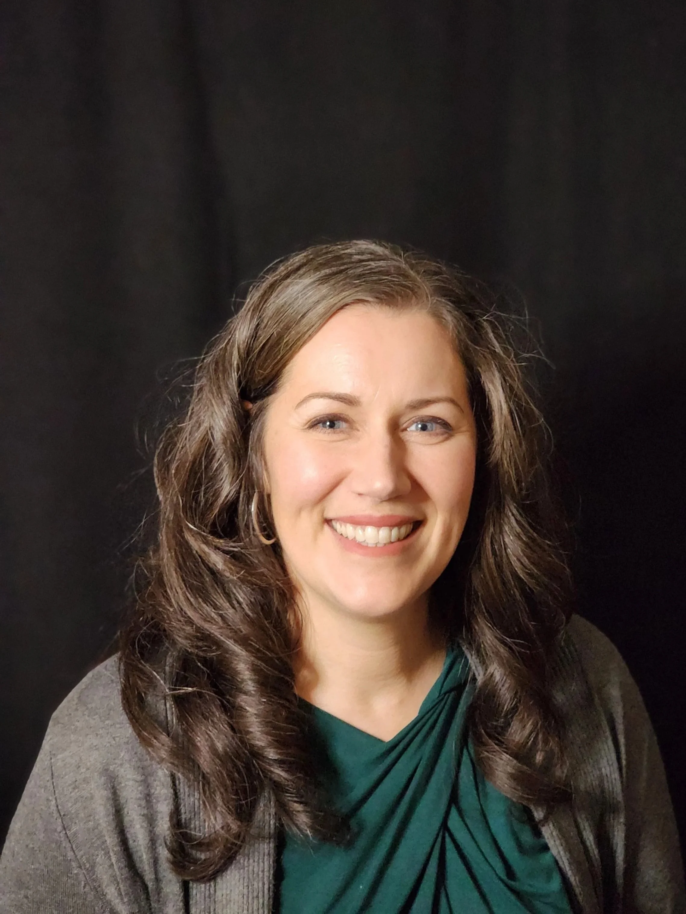 smiling woman with pale skin and long brown hair wearing a gray cardigan and green shirt