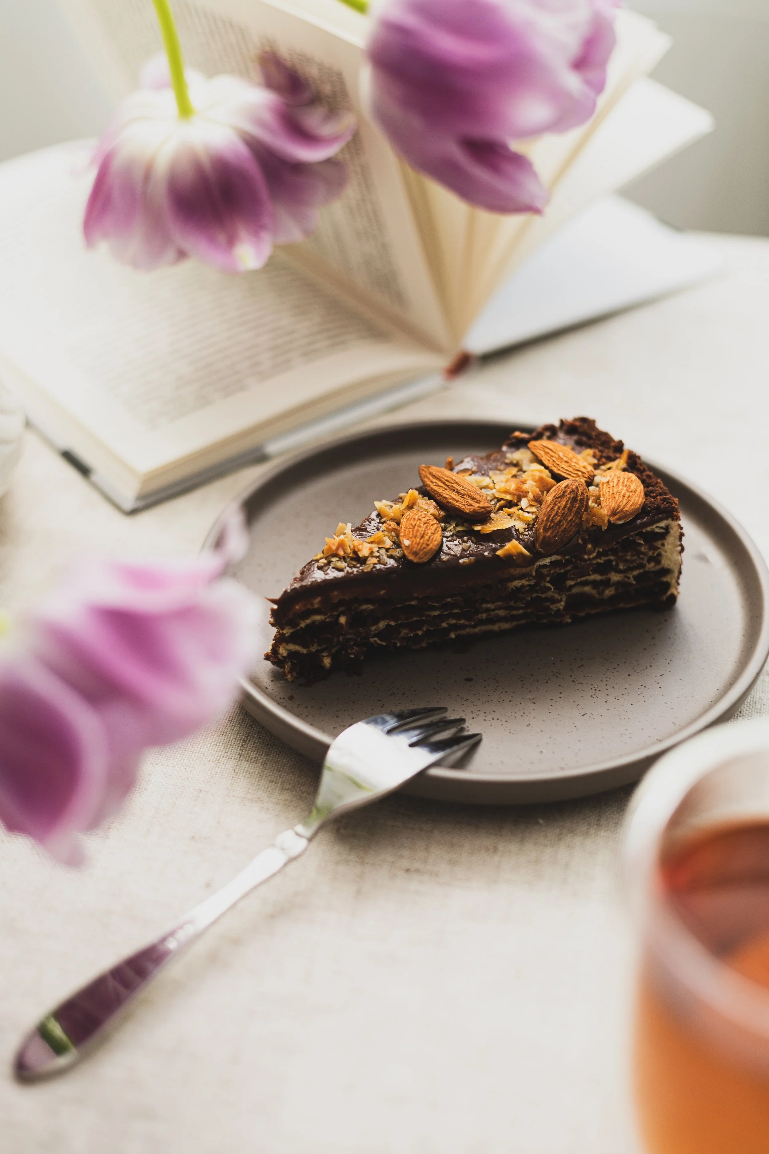A slice of chocolate cake topped with almonds and chopped nuts, placed on a white plate on a table. An open book with blurred purple flowers in the foreground and background.