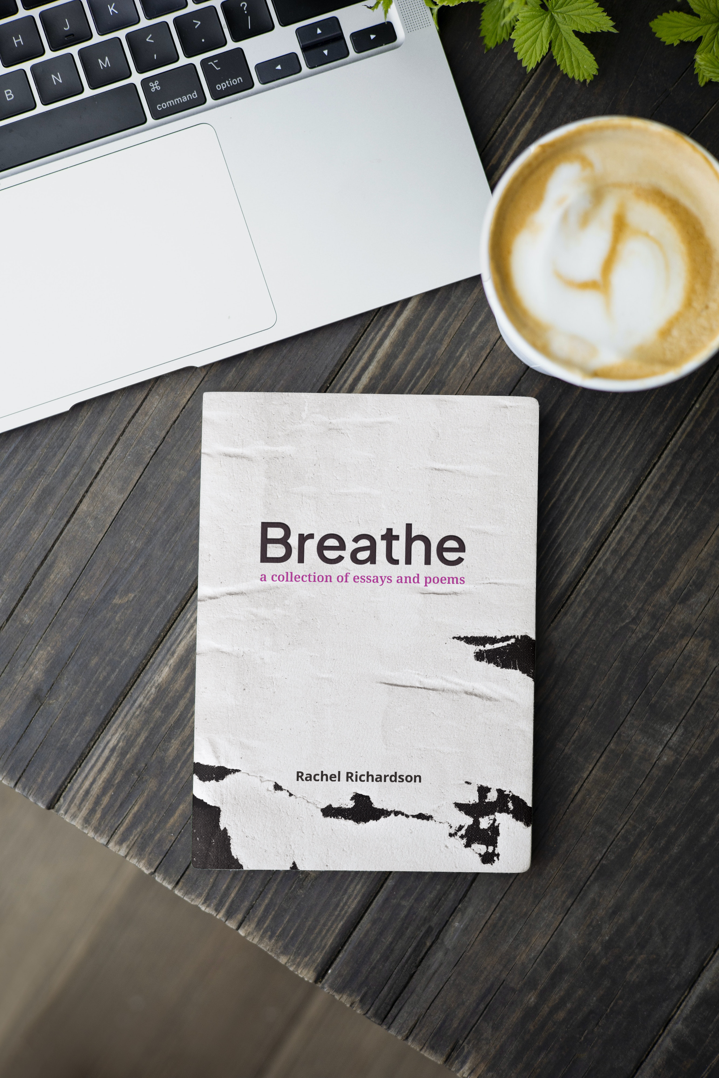 A book titled "Breathe: a collection of essays and poems" by Rachel Richardson on a wooden table, next to a laptop with a black keyboard and a cup of coffee with frothy milk and latte art. Green leaves are partially visible at the top edge.