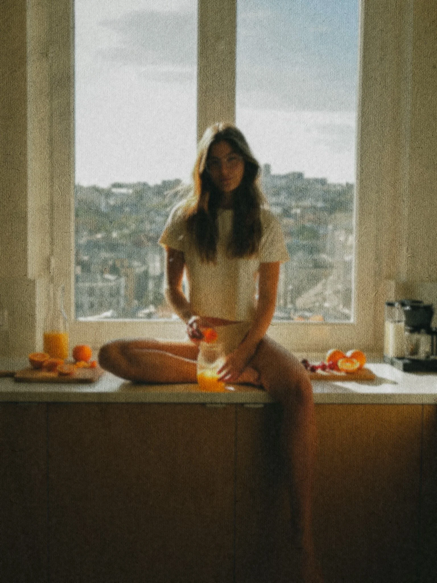 A woman sitting on a kitchen counter with a breakfast spread including juice, oranges, and a coffee maker, with a city view through a large window behind her.