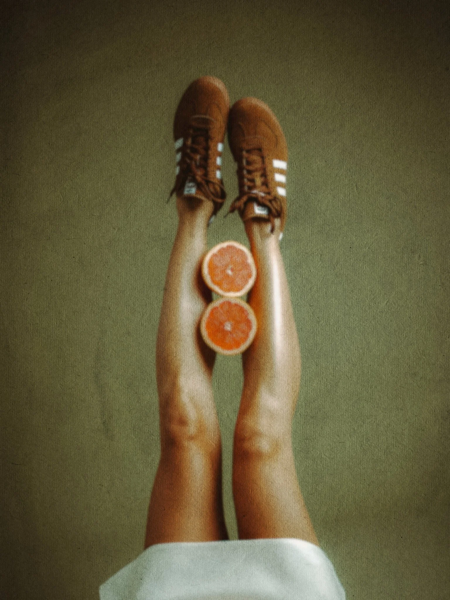 Person lying on their back with legs raised, wearing brown sneakers with white stripes, two grapefruit halves placed between their calves, against a greenish background.
