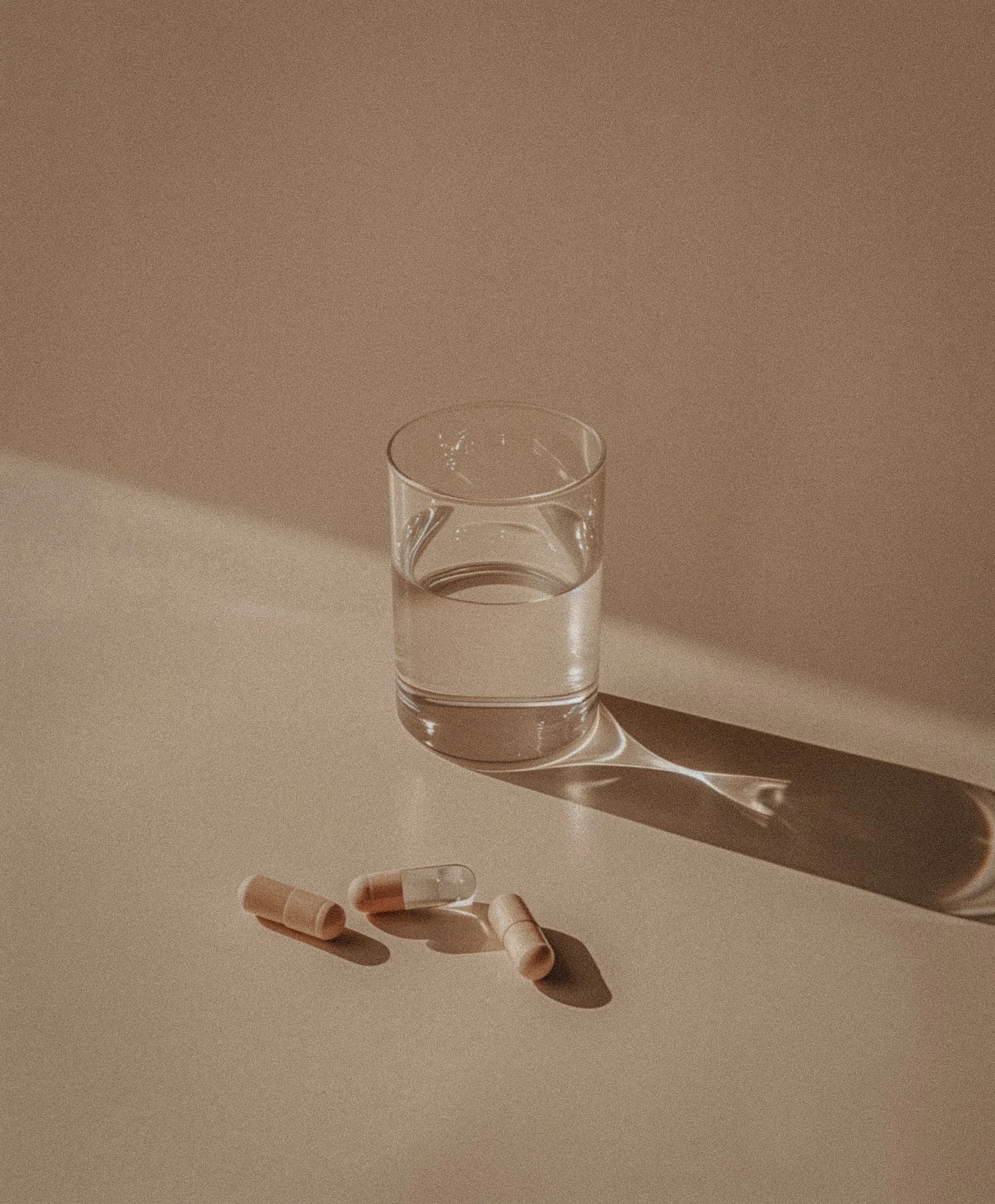 A glass of water and four capsules on a beige surface with shadow and light reflections.